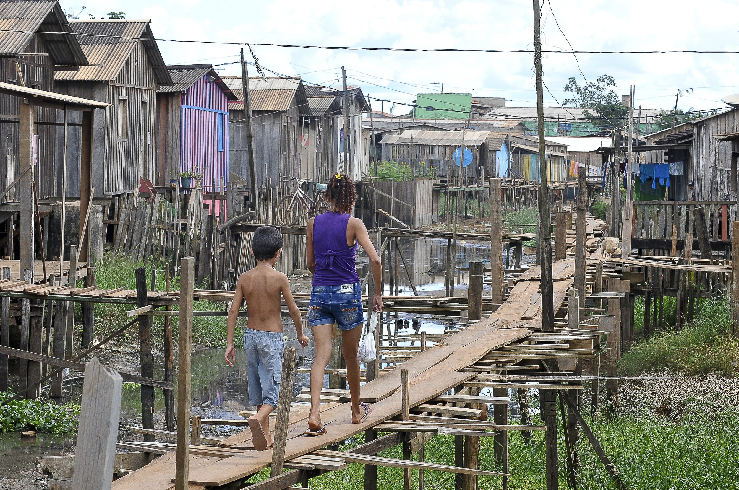 A woman and a young boy walking on a raised wooden plank walkway between rows of stilt houses in a Brazilian palafita village, with weathered wood siding, laundry lines, and standing water beneath.