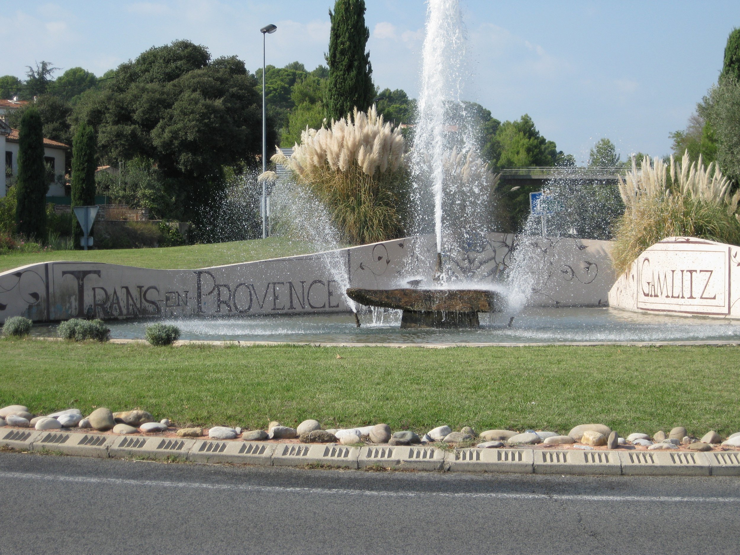 Roundabout at the entrance to the village of Trans-en-Provence in southern France, with a village sign and Provençal landscape in the background.