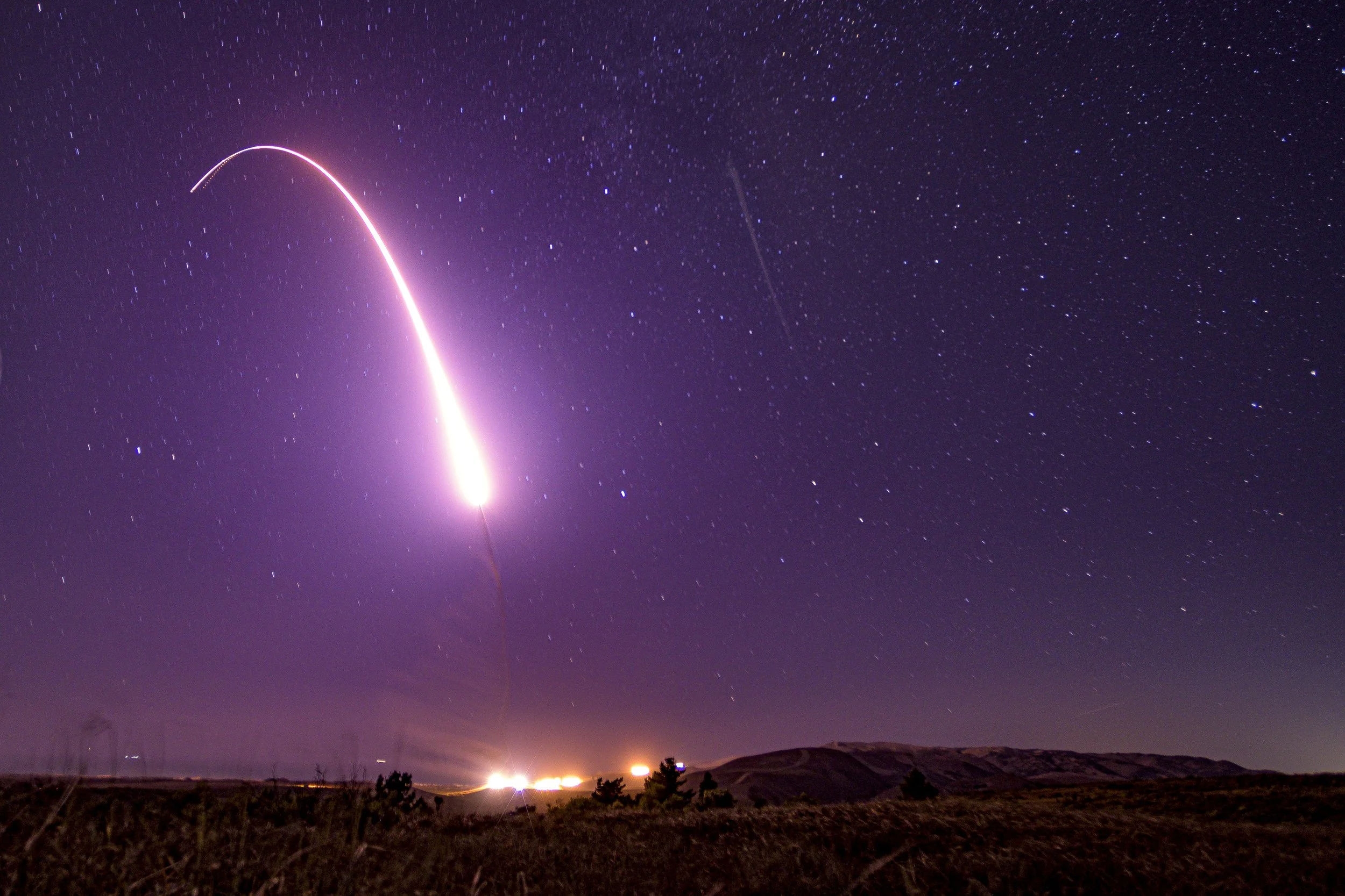 Long-exposure photograph of an unarmed Minuteman III intercontinental ballistic missile launching at night, showing the bright arc of the missile trail rising against a starfield.