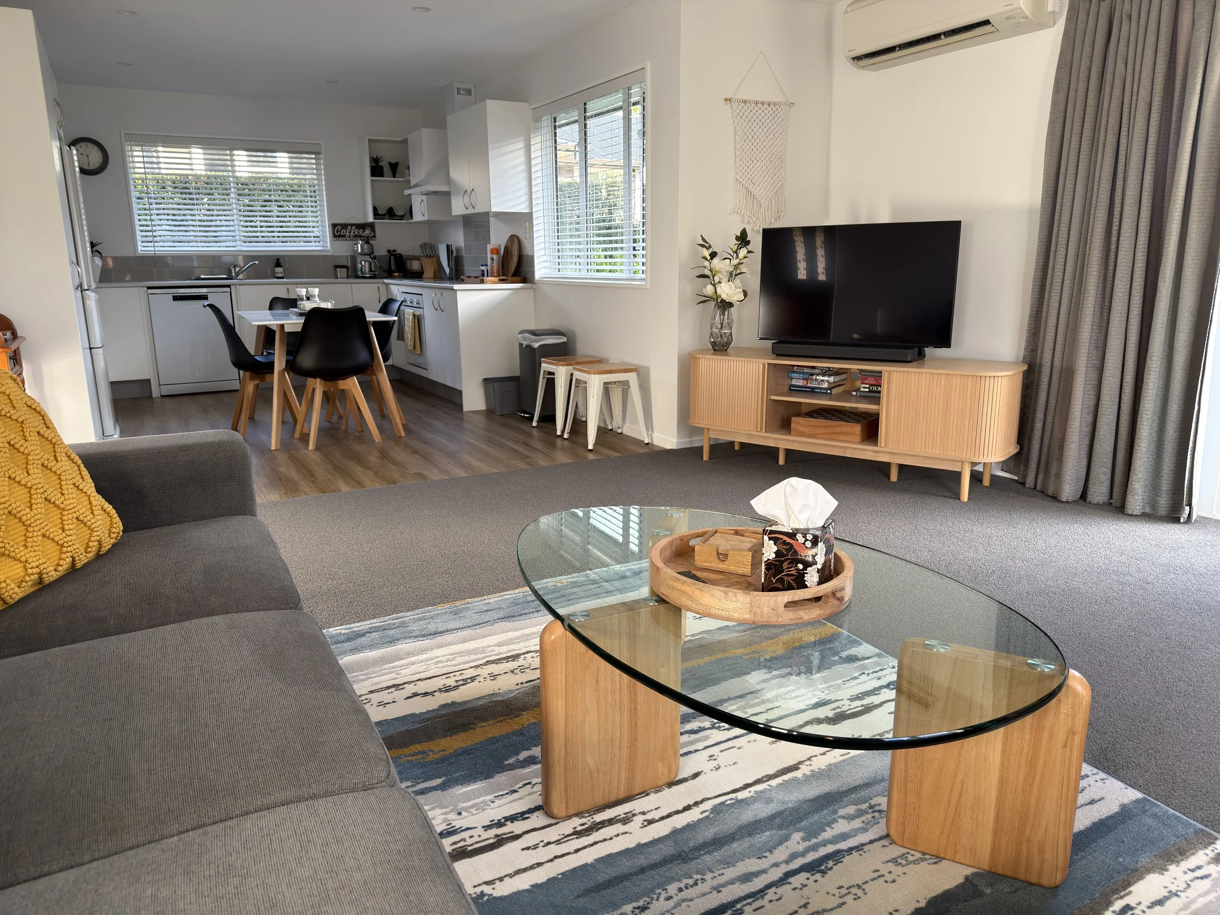 A living room with a gray sofa, a round glass coffee table, a patterned area rug, a wooden TV stand with books, a flat-screen TV, a potted plant, and a macrame wall hanging. The room has windows with white blinds and gray curtains, and a kitchen area