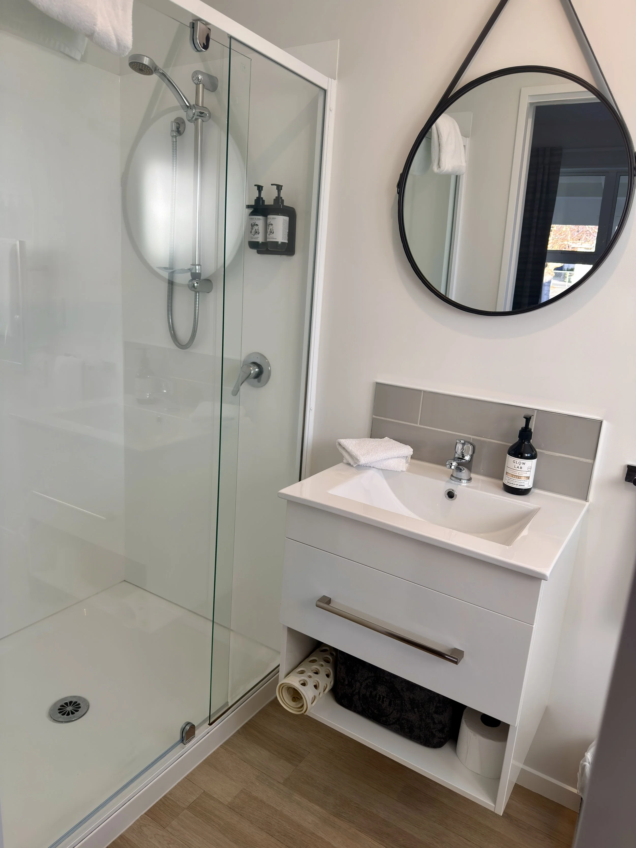 Bathroom with a glass-enclosed shower, a white vanity with a sink, a round mirror hanging above the sink, a towel, and toiletries on the countertop. There are two shelves with wall-mounted soap dispensers inside the shower, a window reflecting outsid
