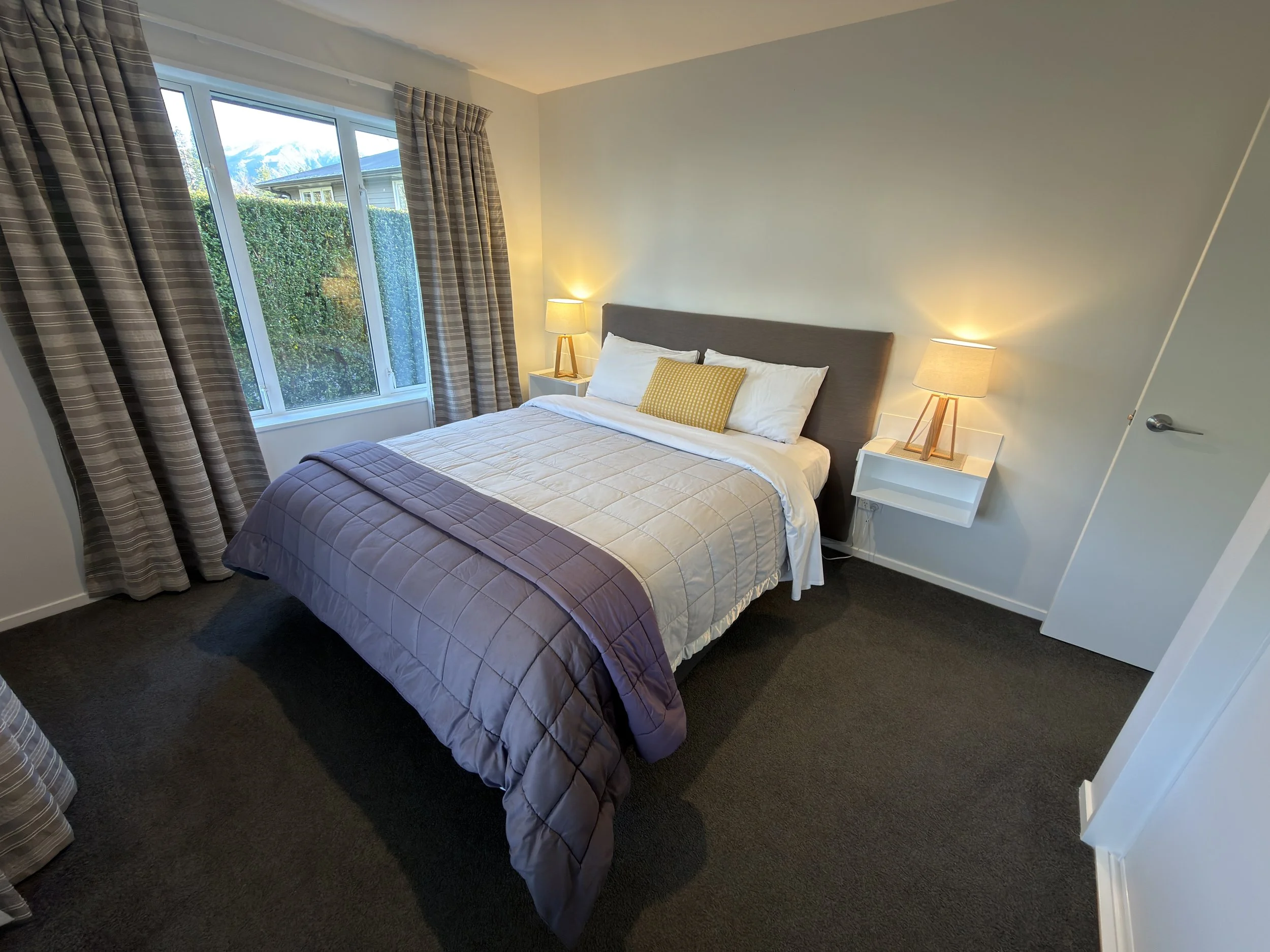 A bedroom with a bed that has a beige quilt and purple bedspread, two white nightstands with lamps, and a window with plaid curtains showing a view of greenery and mountains outside.