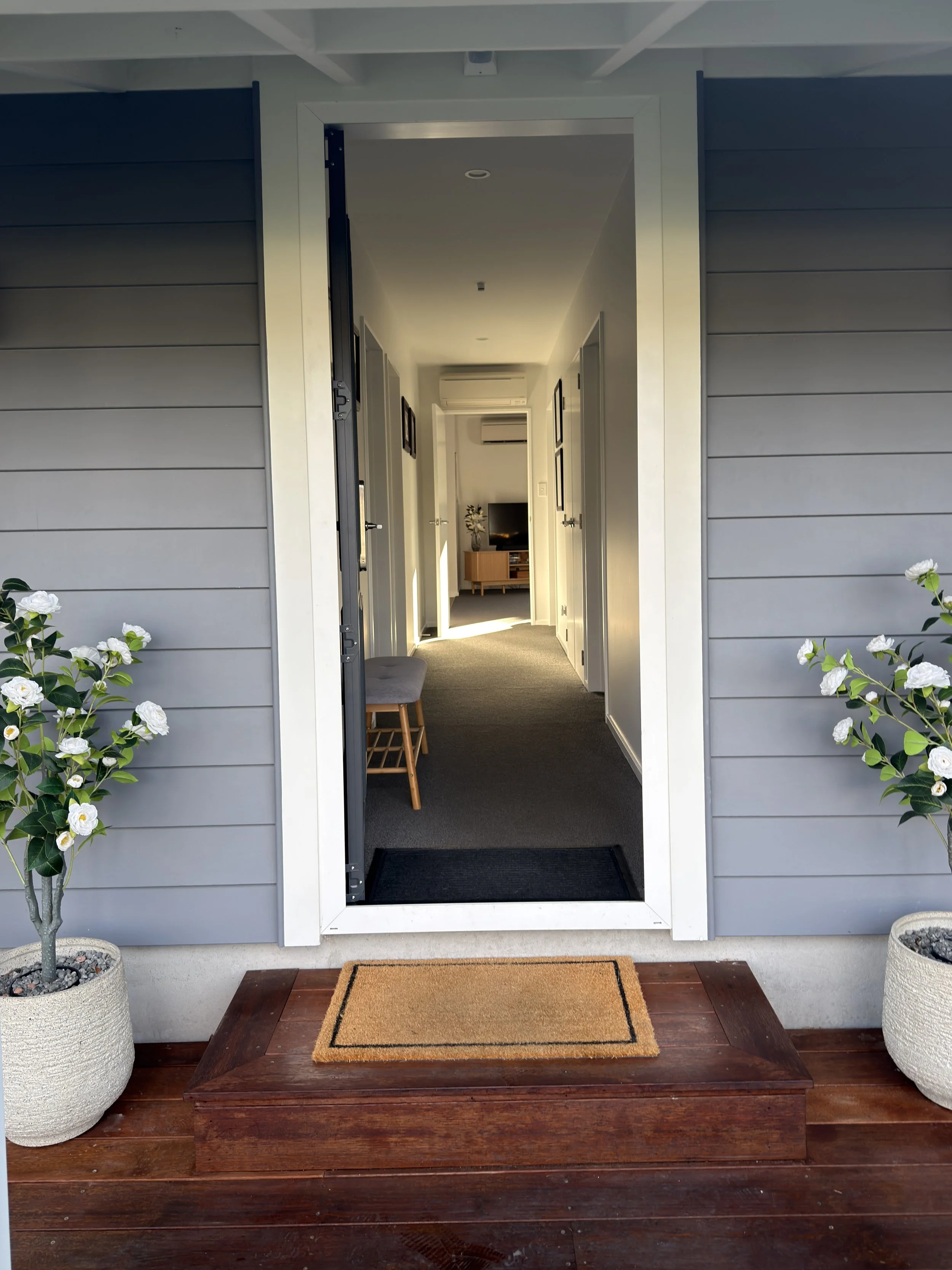 View of an open front door leading into a hallway with a small wooden bench on one side, and a TV and air conditioning unit in the background. The doorframe is white with gray exterior walls, and there are potted plants with white flowers on both sid