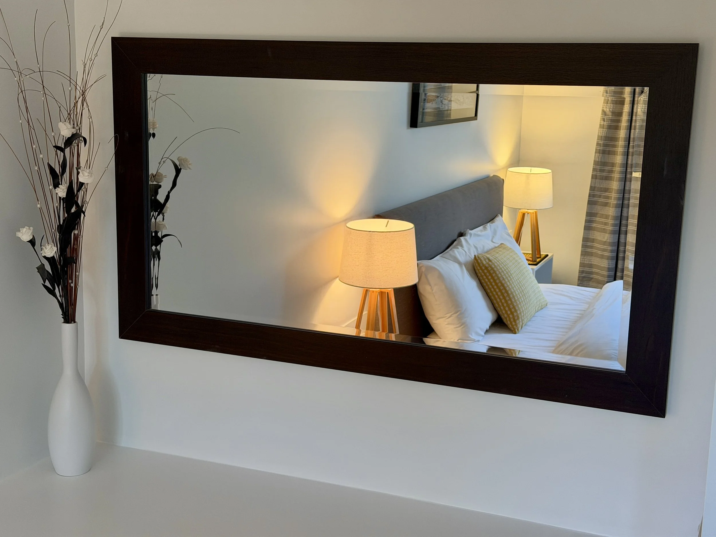 A large mirror with a dark wooden frame reflecting a bedroom with a bed, two lamps on wooden nightstands, and a framed picture on the wall. To the left of the mirror is a white vase with decorative dried branches and white flowers.