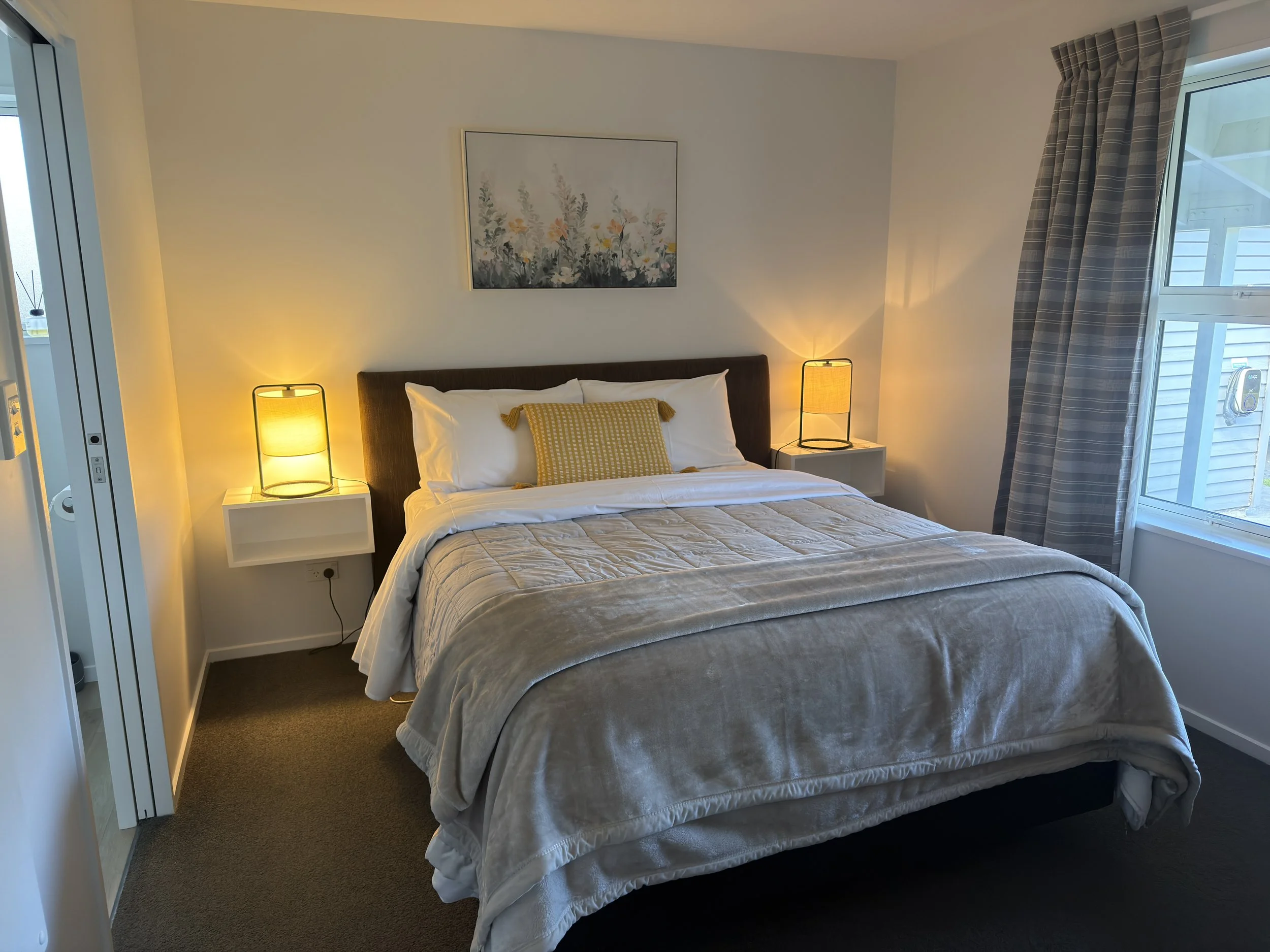 A bedroom with a bed, two hanging bedside lamps, a piece of artwork above the bed, a window with plaid curtains, and a view of an outdoor balcony.