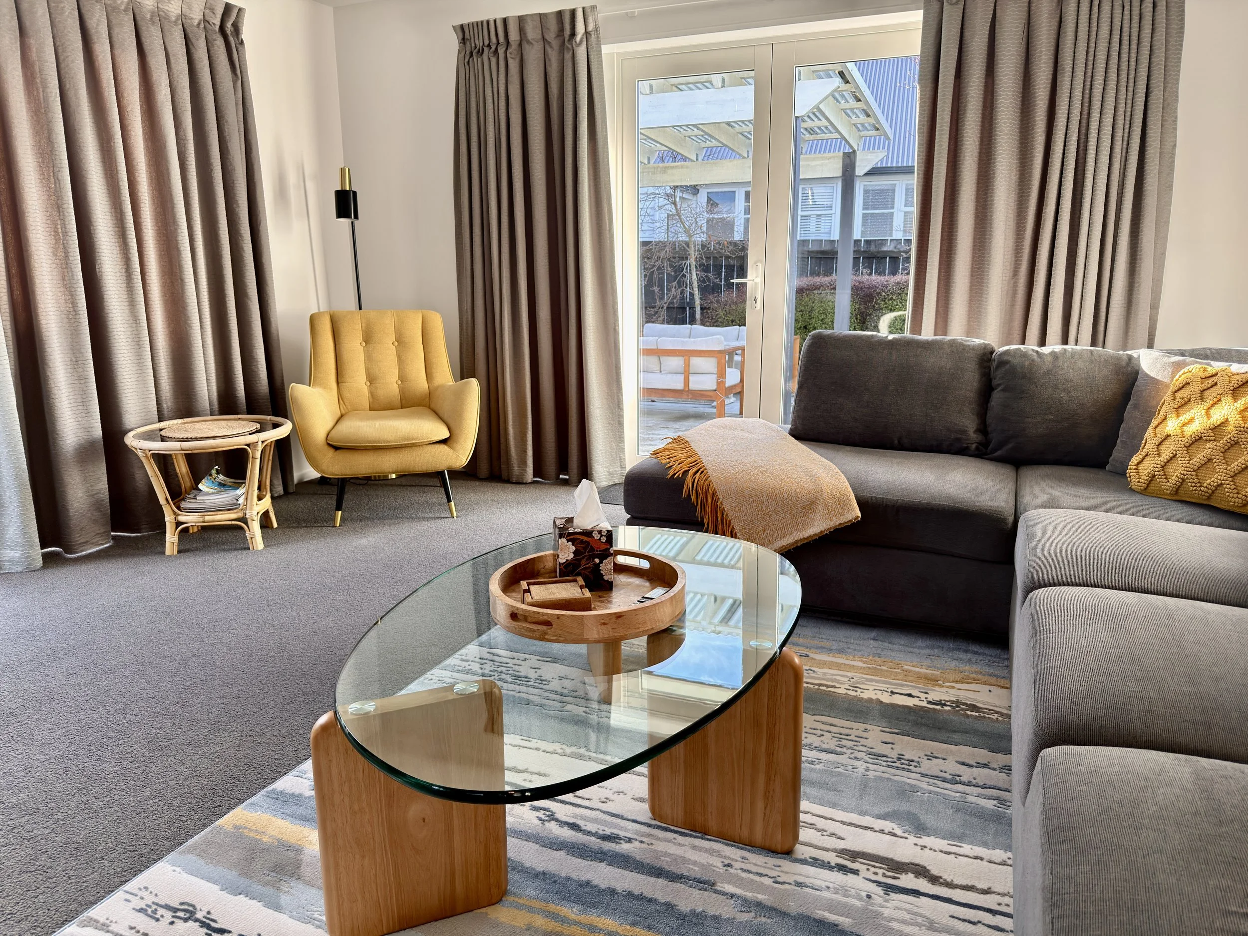 Living room with beige curtains, yellow armchair, gray sofa, glass coffee table, and sliding door leading to outdoor patio with white furniture and wooden fencing.