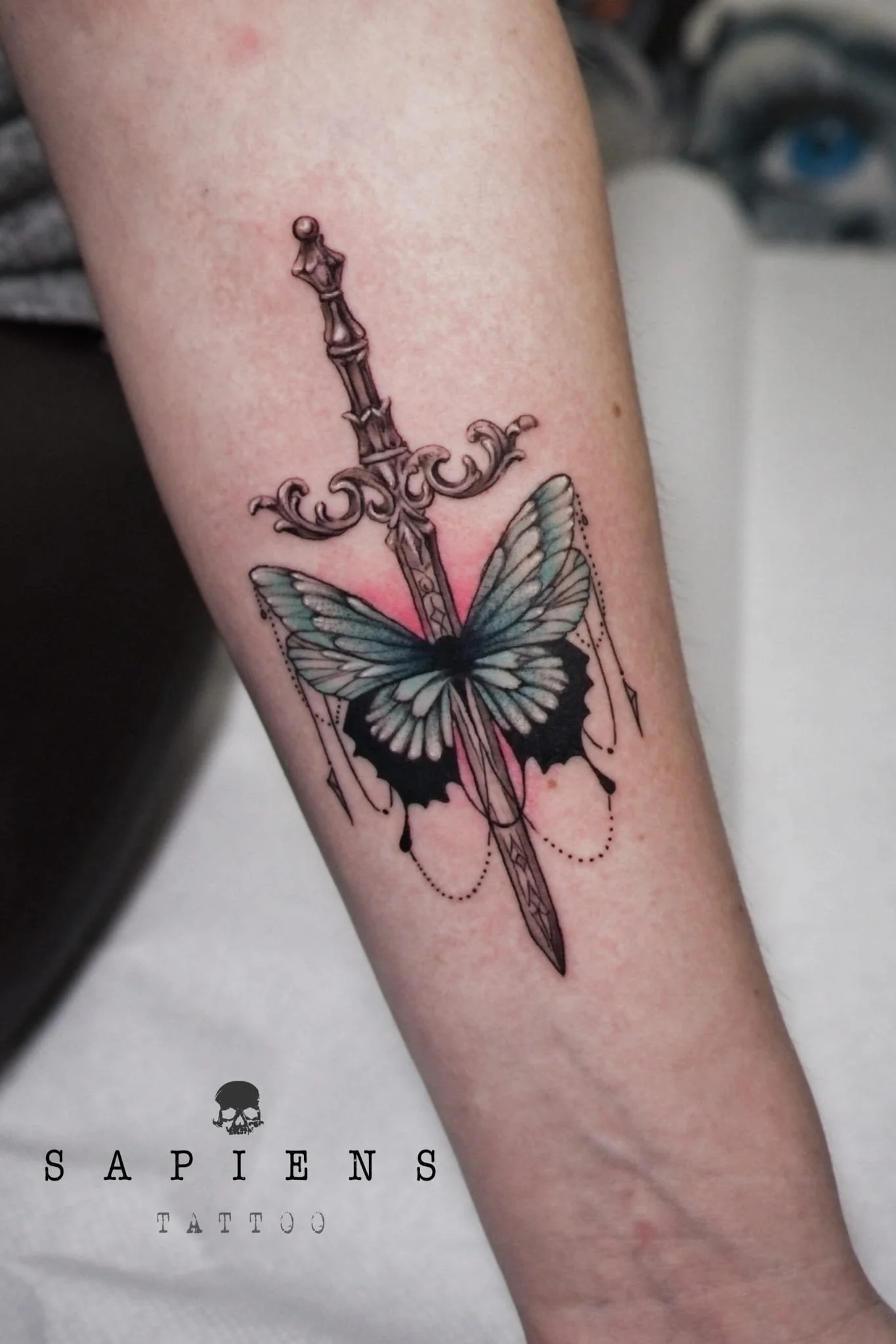 tatuaj pe antebrat cu fluture si aripi colorate cu verde deschis si o sabie realistica