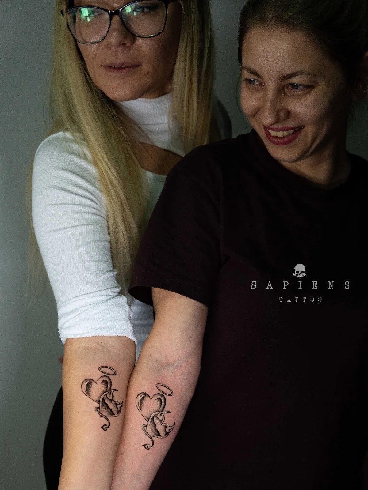 doua femei cu un tatuaj matching realizat pe antebrat interior cu doua inimioare una simbolizand un dracusor si cealalta un inger
