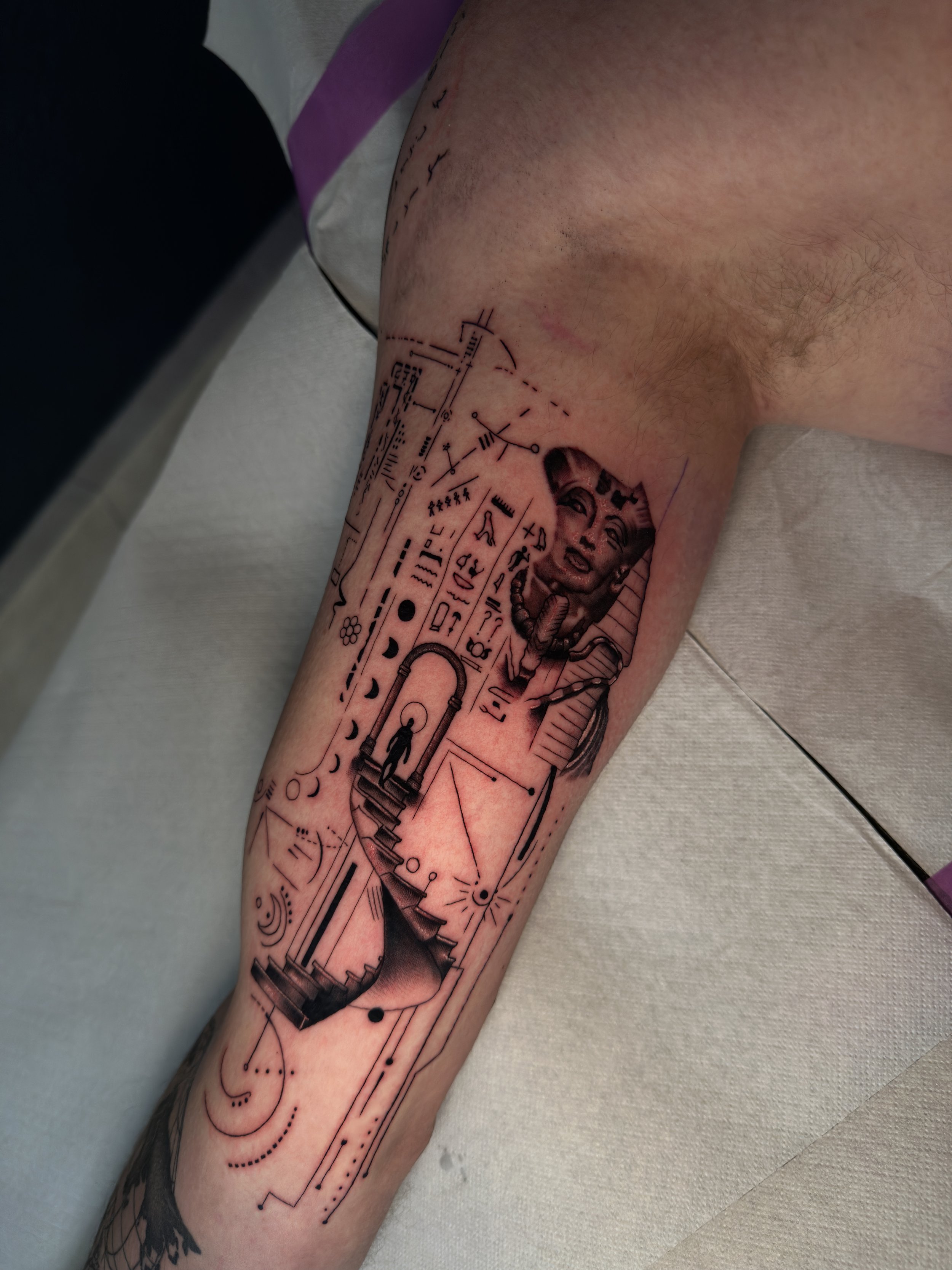 Tattoo auf einem Unterarm, das geometrische und technische Elemente, eine Frauenkopfzeichnung, eine Treppe und verschiedene Symbole enthält.