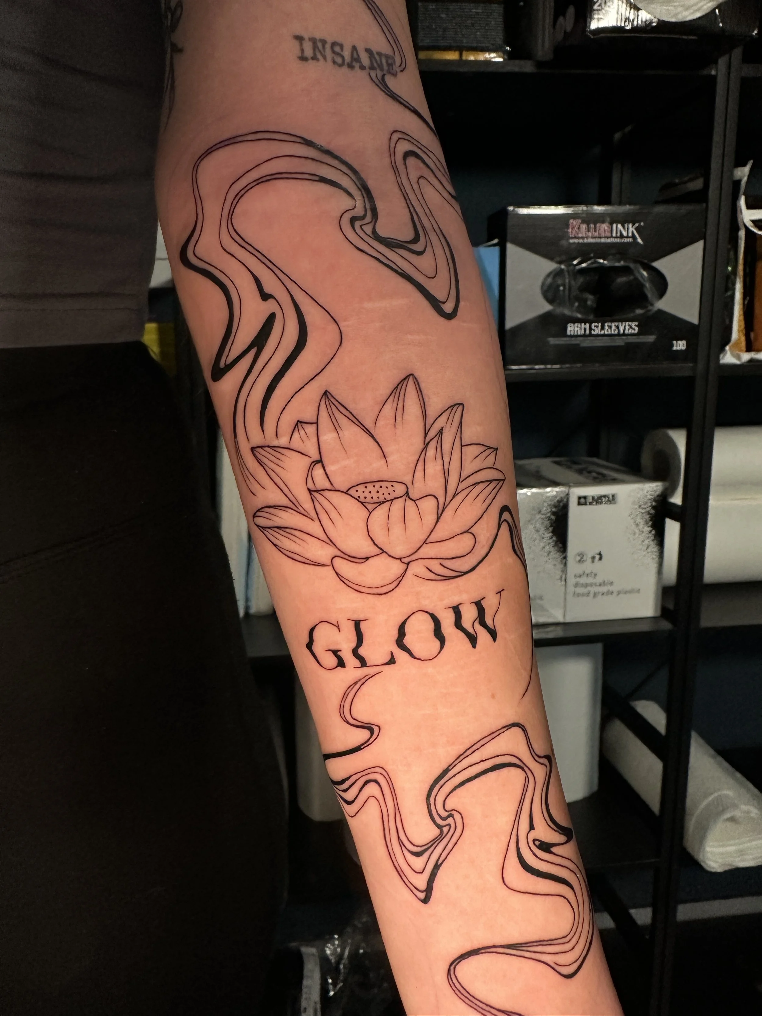 Tattoo eines stilisierten Seerosens, umgeben von abstrakten Linien, mit dem Wort 'GLOW' darunter.