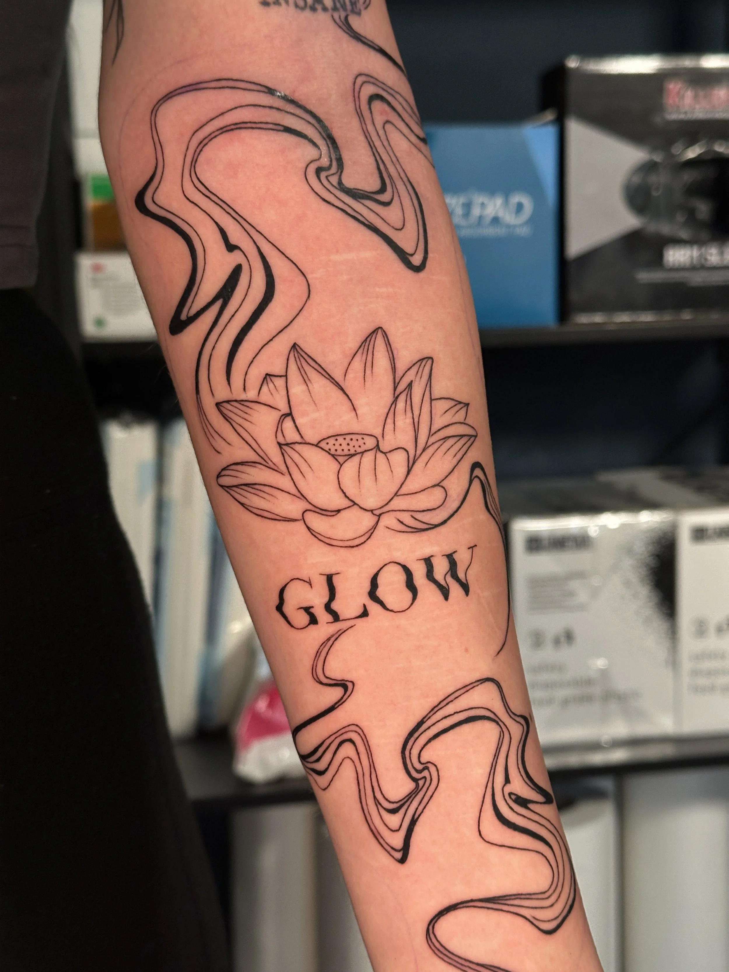 Tattoo eines Lotusblume mit Rauchschlangen und dem Wort »GLOW« auf dem Unterarm.