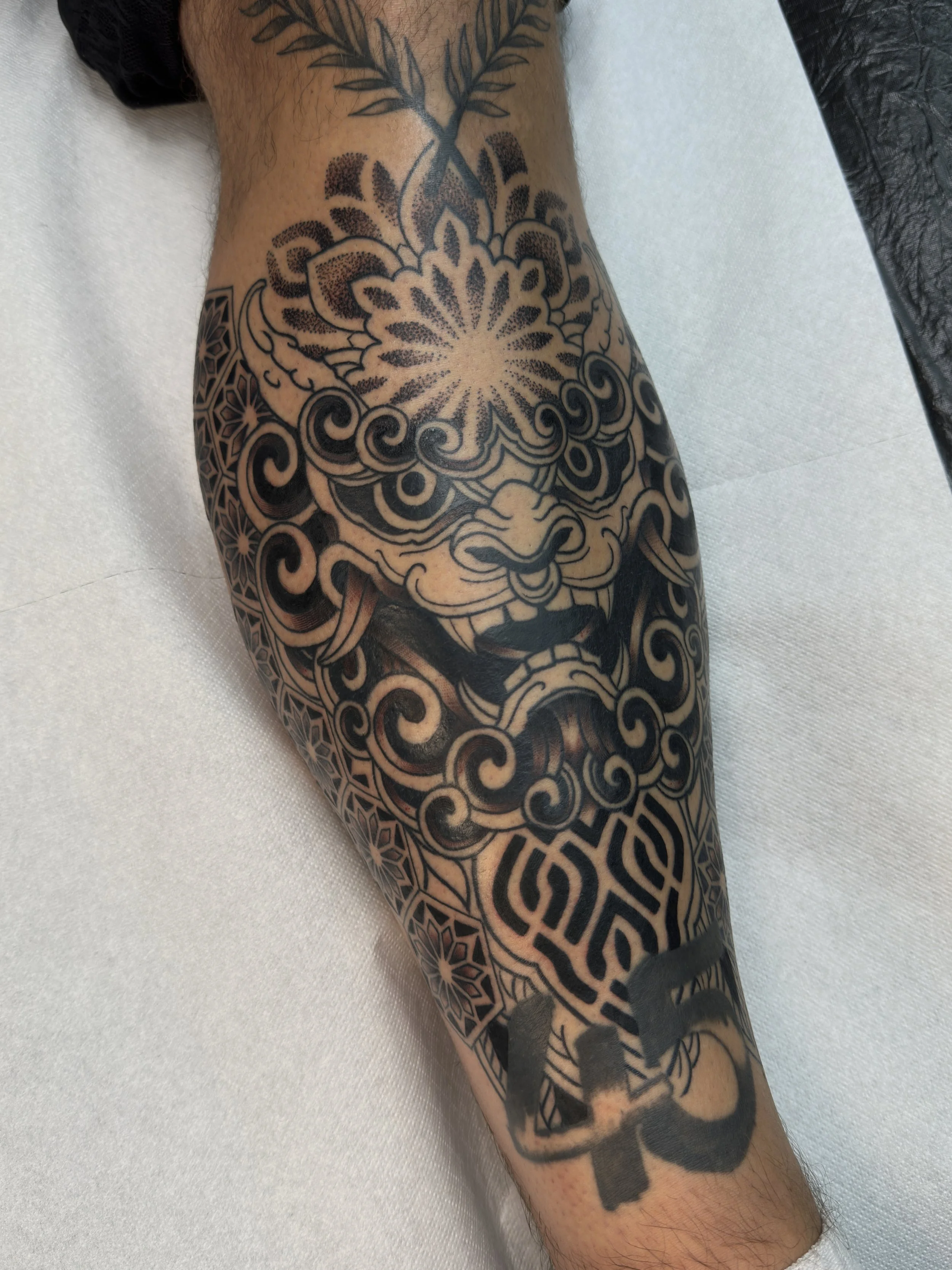 Schwarze Traditional-Tattoo auf einem Unterarm mit Löwenkopf, geometrischen und floral-inspirierten Mustern, und schwarzen Buchstaben 'CH'.