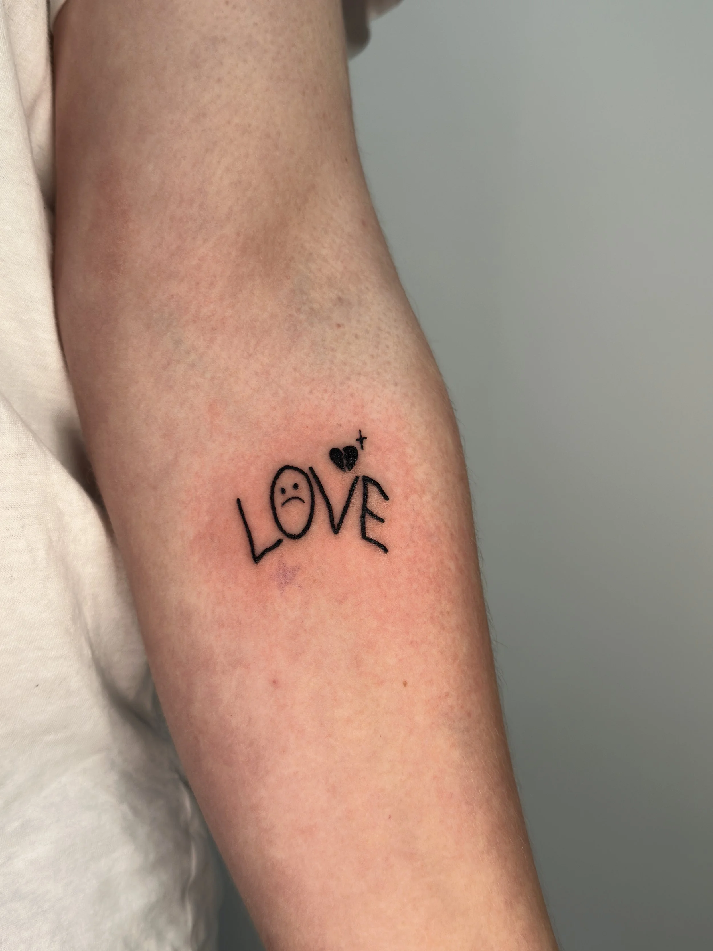 Tattoo auf dem Oberarm mit dem Wort 'LOVE' und einem traurigen Gesicht als 'O', einem schwarzen Herz und einem Pluszeichen.
