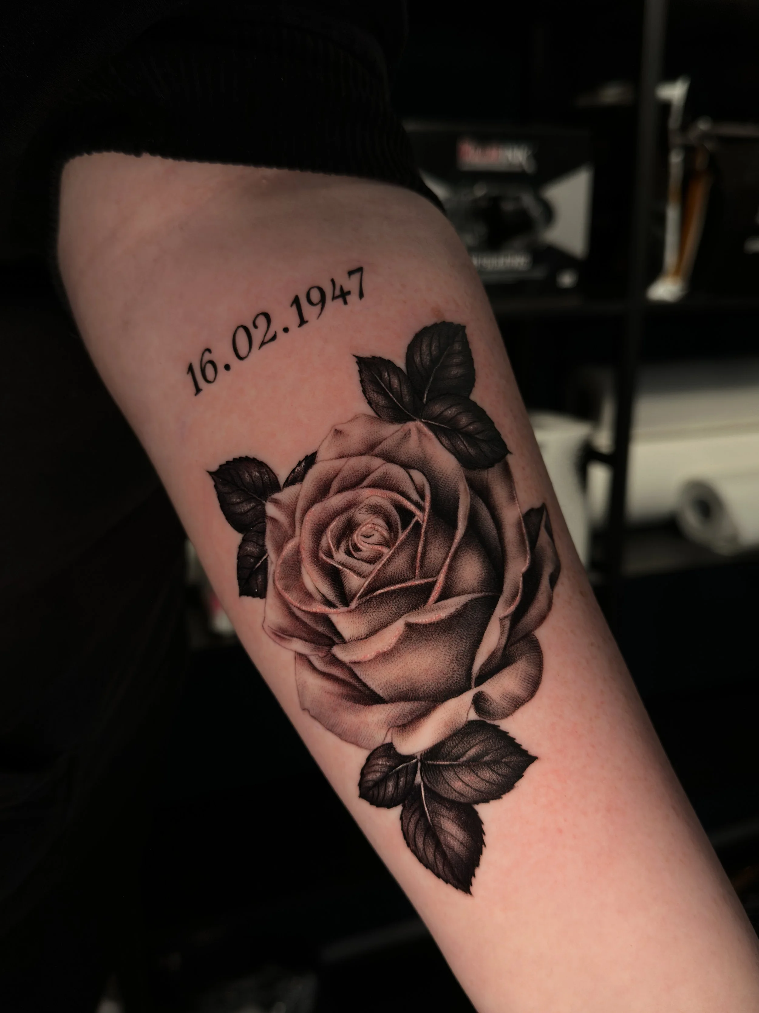 Tattoo einer Rose mit Blättern und einem Datum 16.02.1947 auf dem Oberarm.