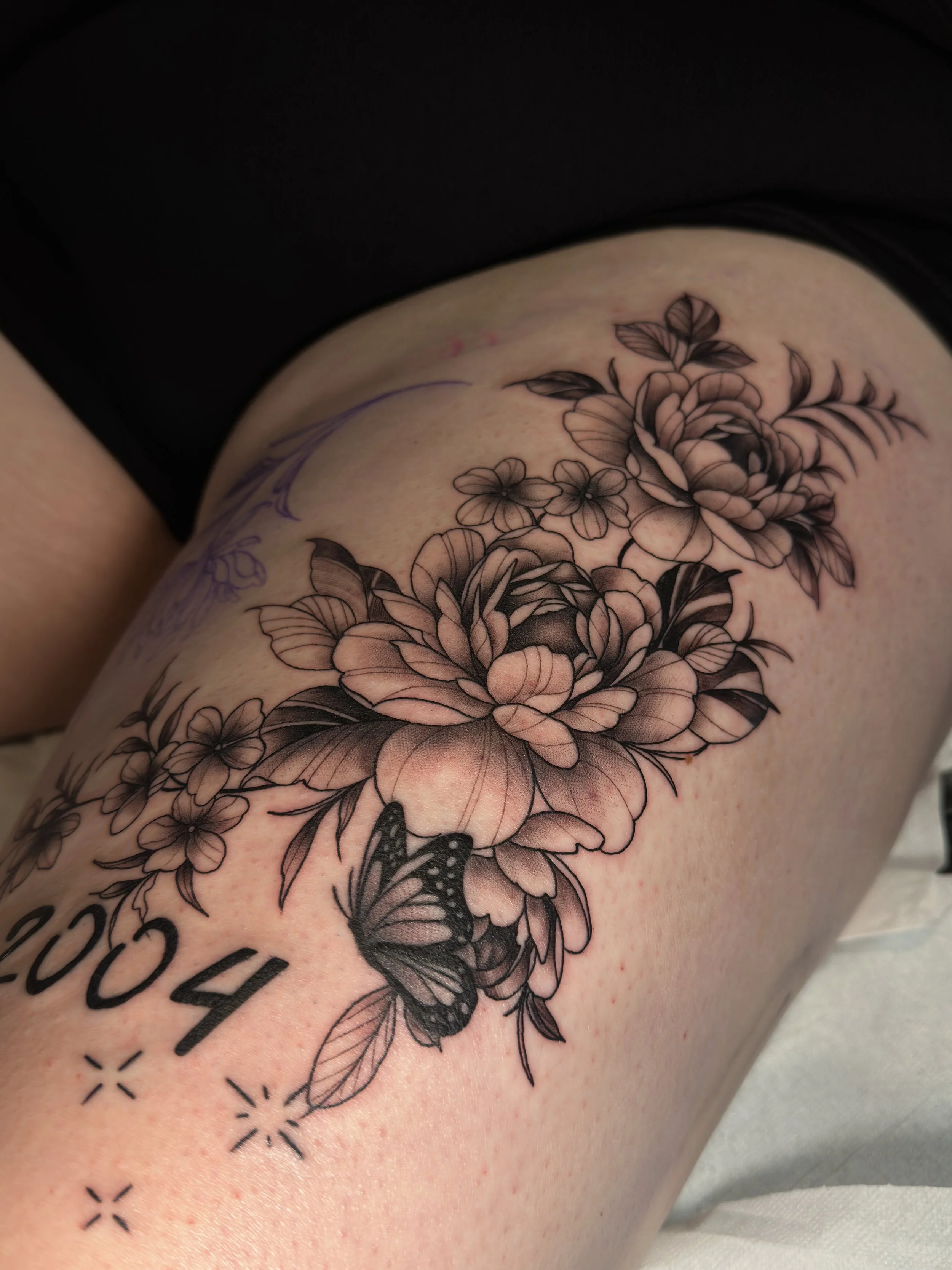 Schwarze und graue Tattoos von Blumen und einem Schmetterling am Oberschenkel.