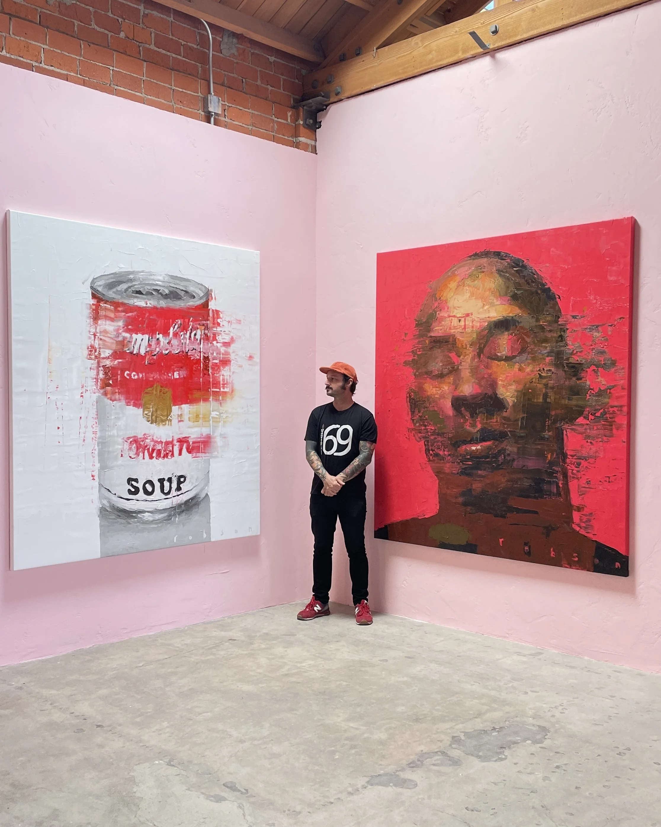 An art gallery with pink walls and two large paintings; one of a soup can and one of a woman with glasses. A man with tattoos, wearing a black shirt, red sneakers, and an orange cap, stands in front of the paintings.