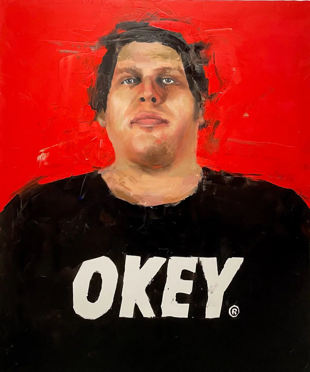 A portrait of a person with black hair, wearing a black shirt with the word "OKEY" in white letters, set against a bright red background.