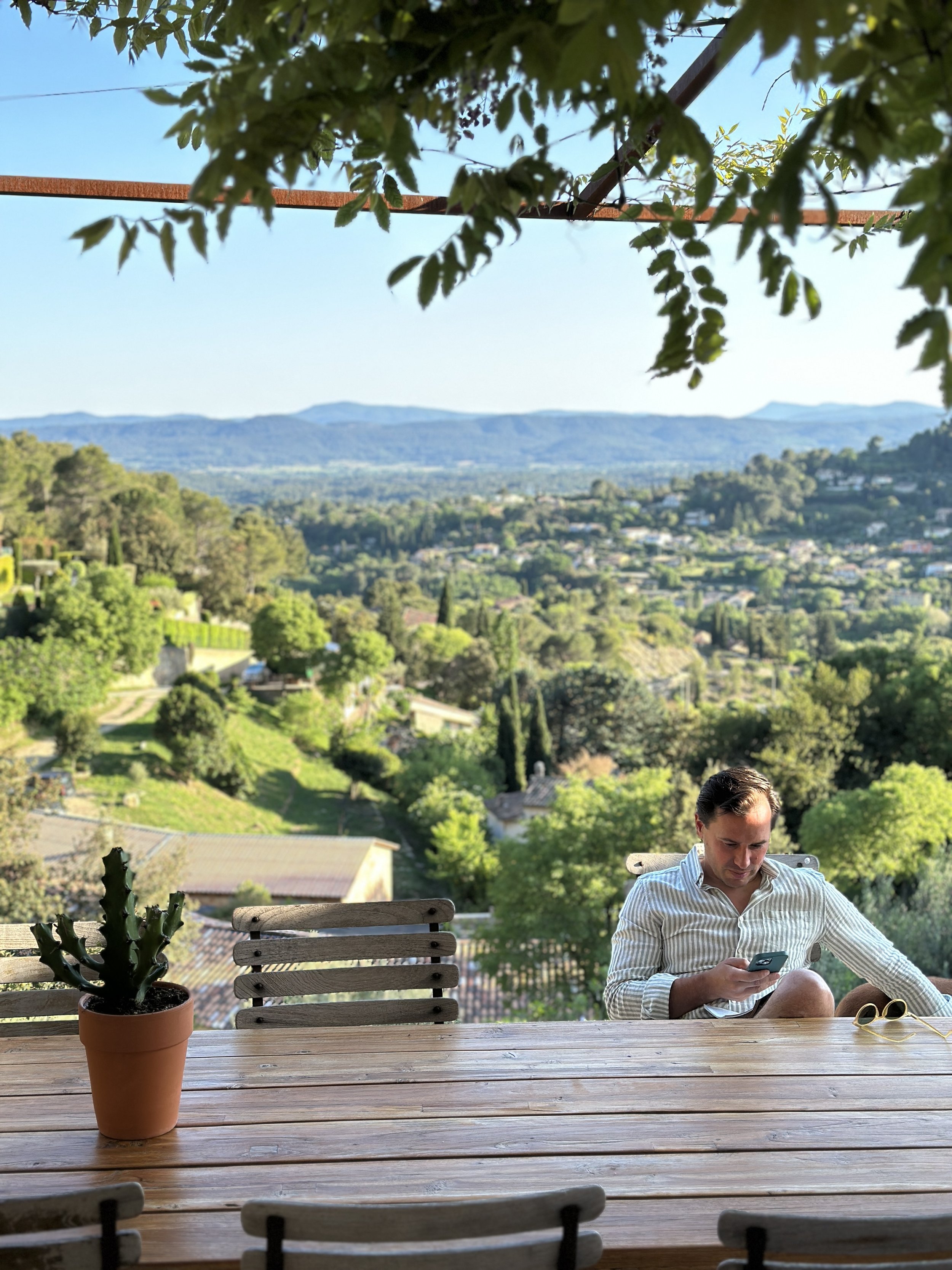 A man sitting at an outdoor wooden table, looking at his phone, with a scenic green valley and mountains in the background. Potted cactus on the table, with leafy branches above. It appears to be late afternoon.