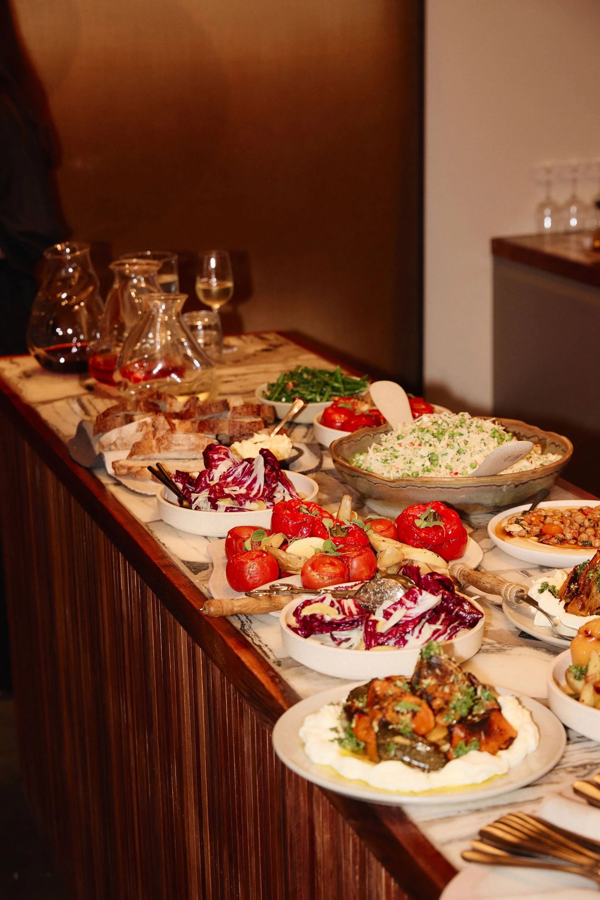 A buffet table filled with various dishes including salads, roasted vegetables, bread, and drinks, set in an indoor dining area.