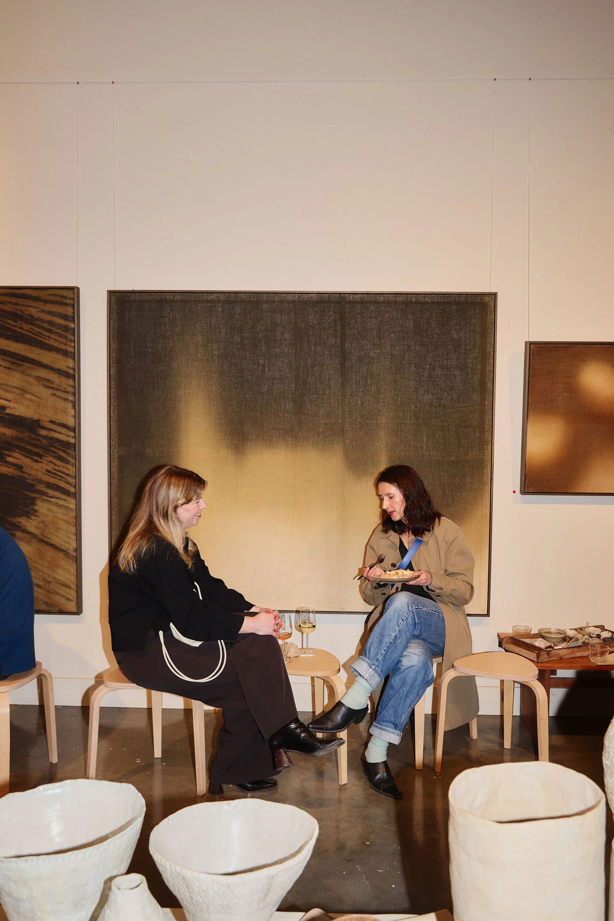 Two women sitting on stools and chatting at an art gallery or museum. They are surrounded by modern artwork on the walls and empty ceramic vases in the foreground.