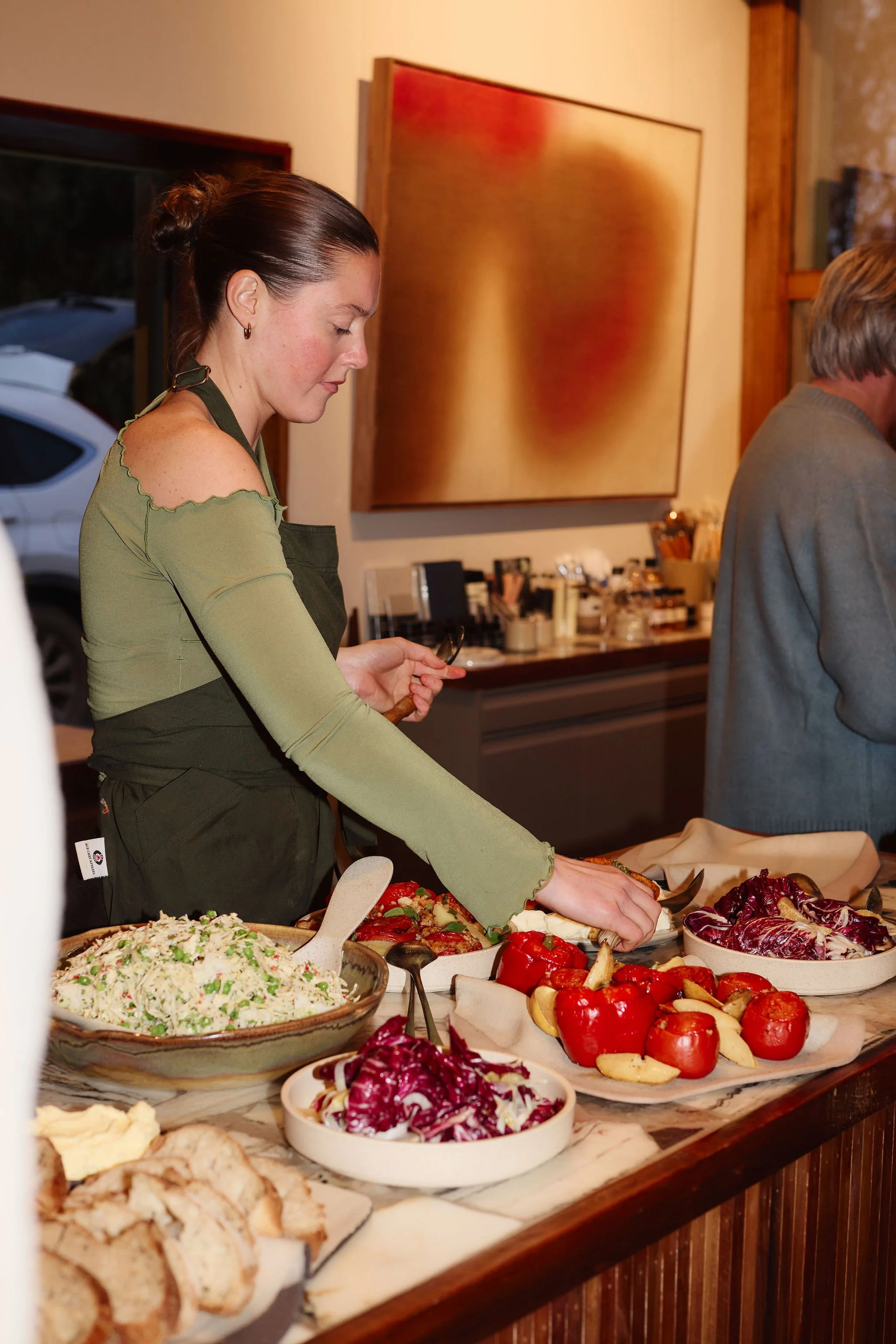A woman wearing a green top and apron is serving food at a buffet dinner table with various salads, vegetables, and dishes. The background shows a framed abstract painting and a wood-paneled space.
