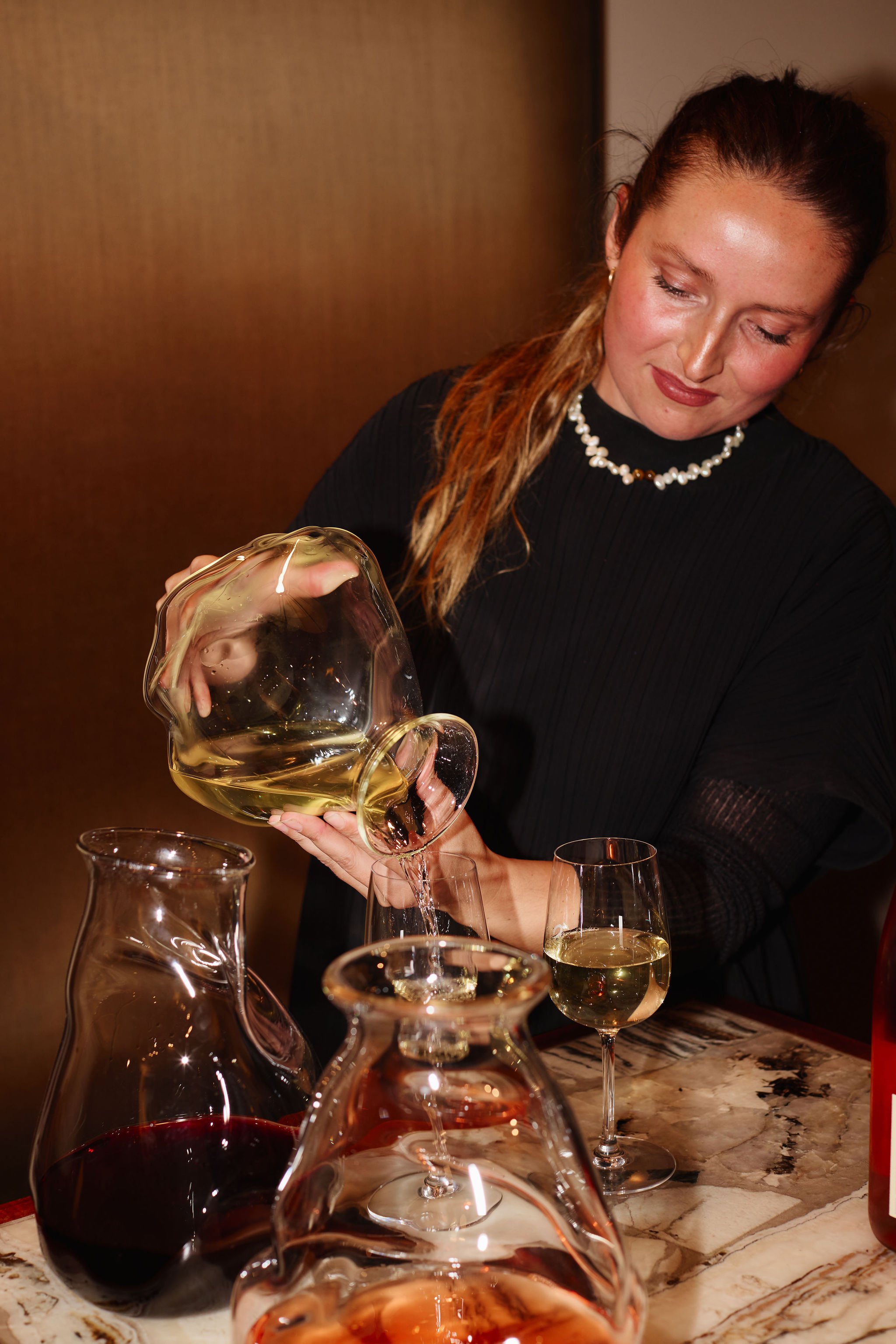 A woman pouring white wine into a glass at a marble table, with multiple wine glasses and decanters filled with red, white, and rosé wine around her.