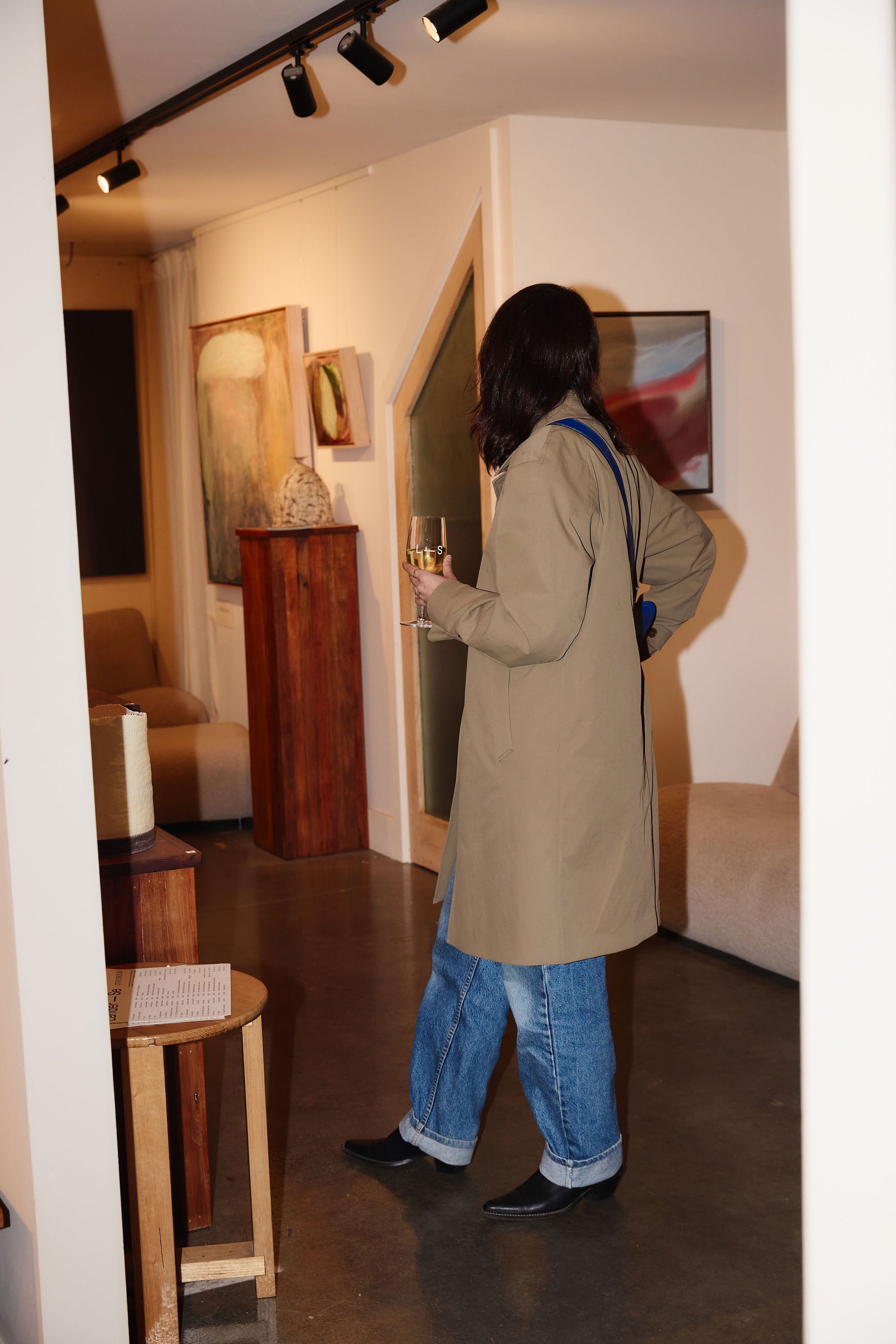 A woman with black hair in a beige trench coat, blue jeans, and black shoes, holding a glass of white wine, stands in an art gallery looking at a painting on the wall.