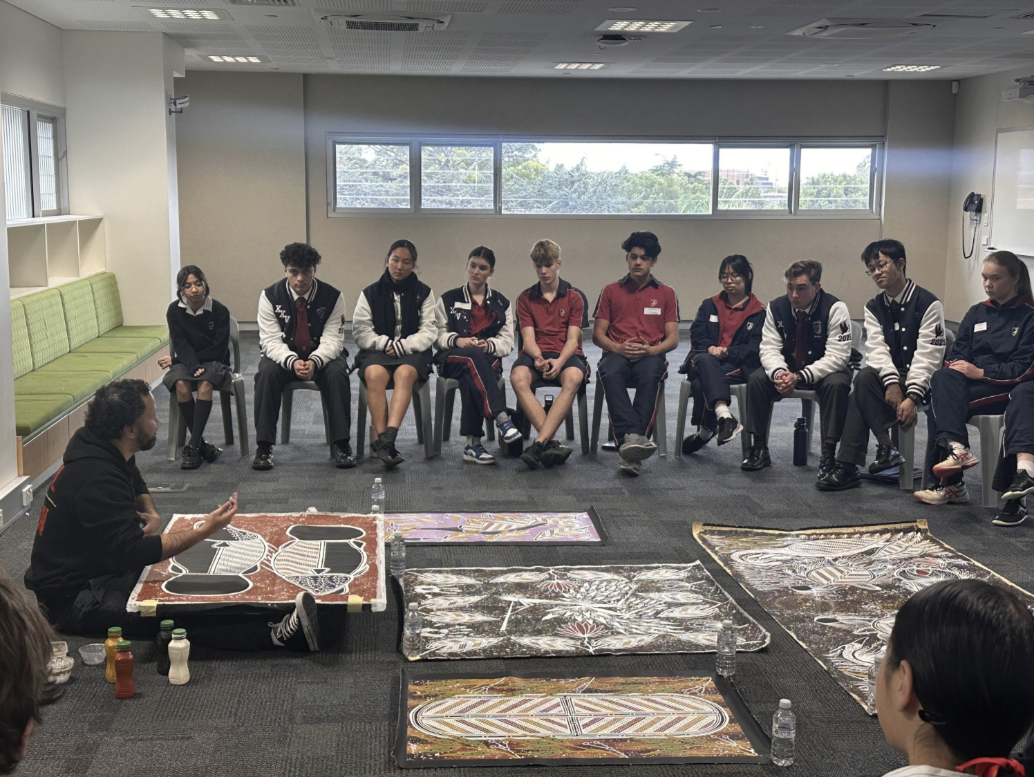 A group of students sitting in a circle in a classroom, listening to a person sitting on the floor who appears to be leading a discussion or presentation, with artwork displayed on the floor in front of them.