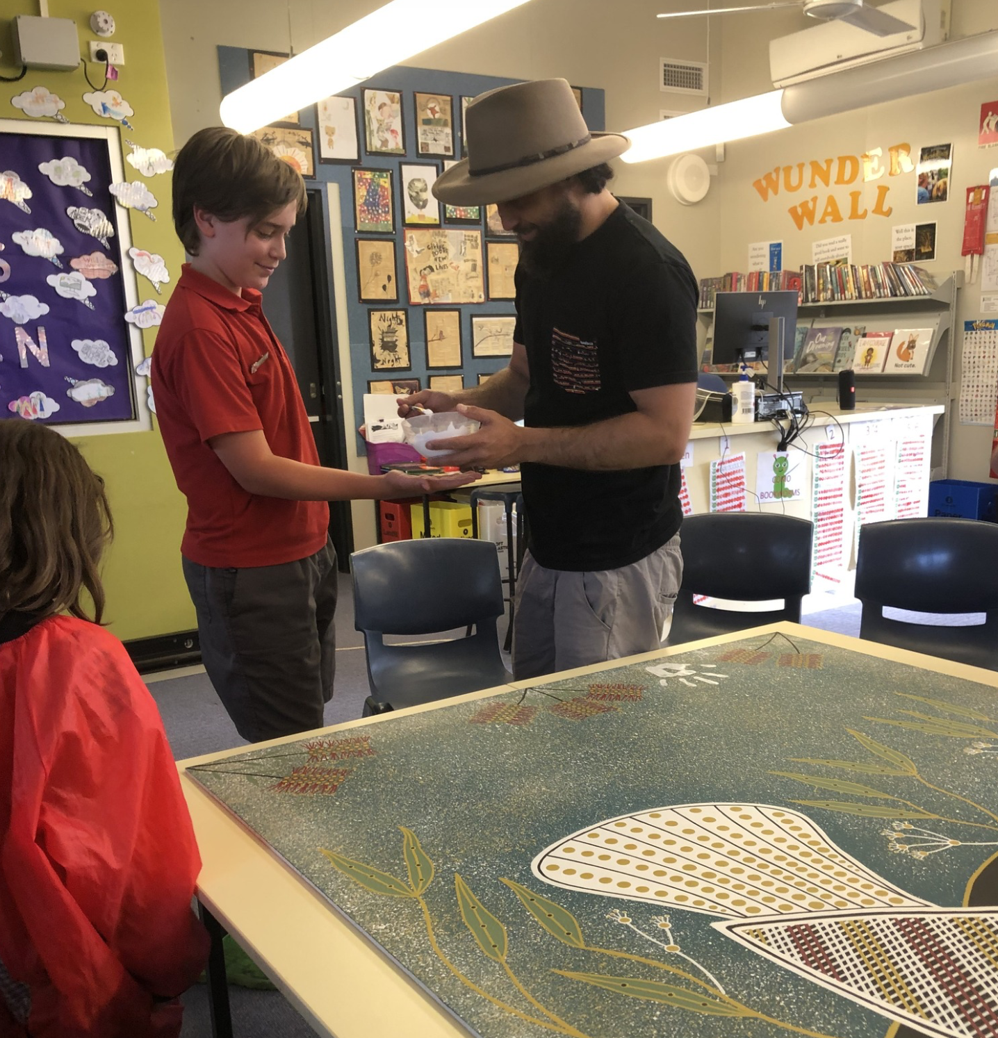 A man wearing a wide-brimmed hat and black t-shirt hands an object to a young boy in a red shirt inside a classroom. Two other children are partially visible, one seated and wearing a red jacket. The classroom has colorful artwork, a decorated wall with clouds, and a counter with books and a sign that reads 'Wunder Wall'.