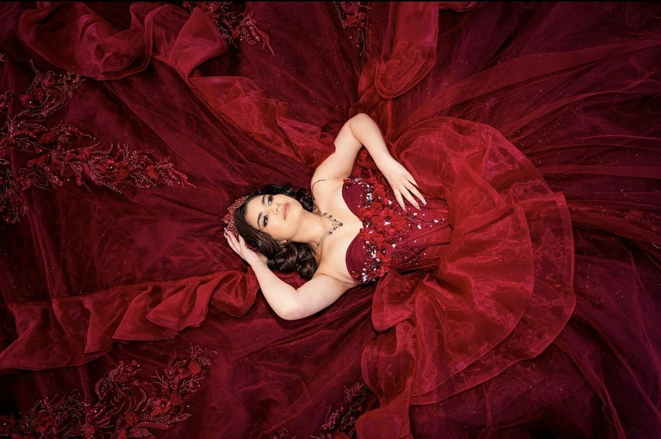 A woman lying on her back surrounded by a large, flowing red dress with embroidery and sequins, looking at the camera with one hand on her head and the other resting on her stomach.