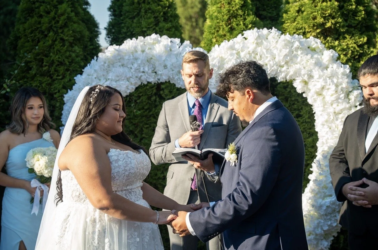 Bride and groom holding hands during a wedding ceremony outside, with officiant reading from a book, surrounded by bridesmaids and groomsmen, with a floral arch and greenery in background.