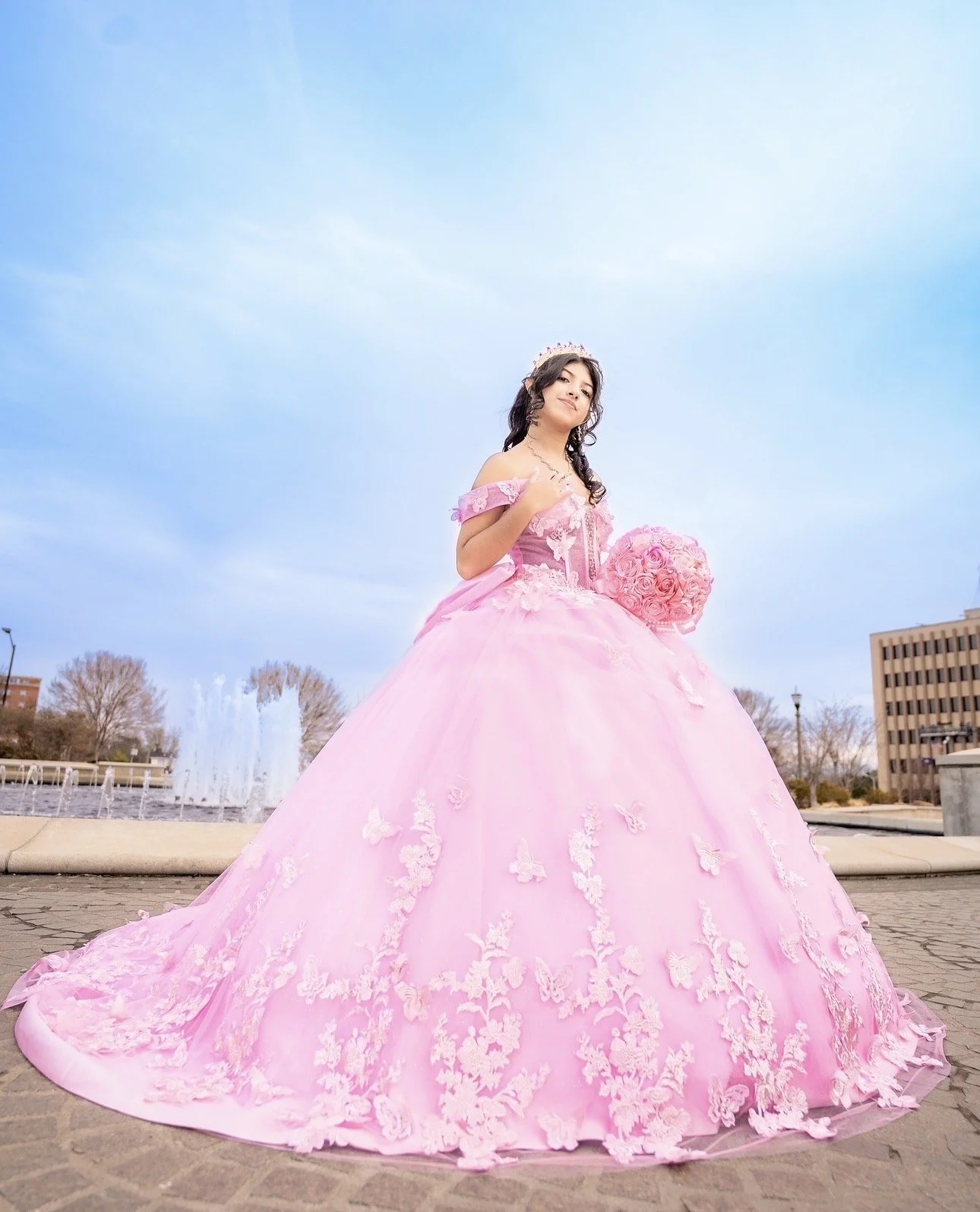 A young woman wearing a pink princess-style gown with floral embroidery, holding a bouquet of pink roses, standing outdoors in a city park with fountains and buildings in the background.