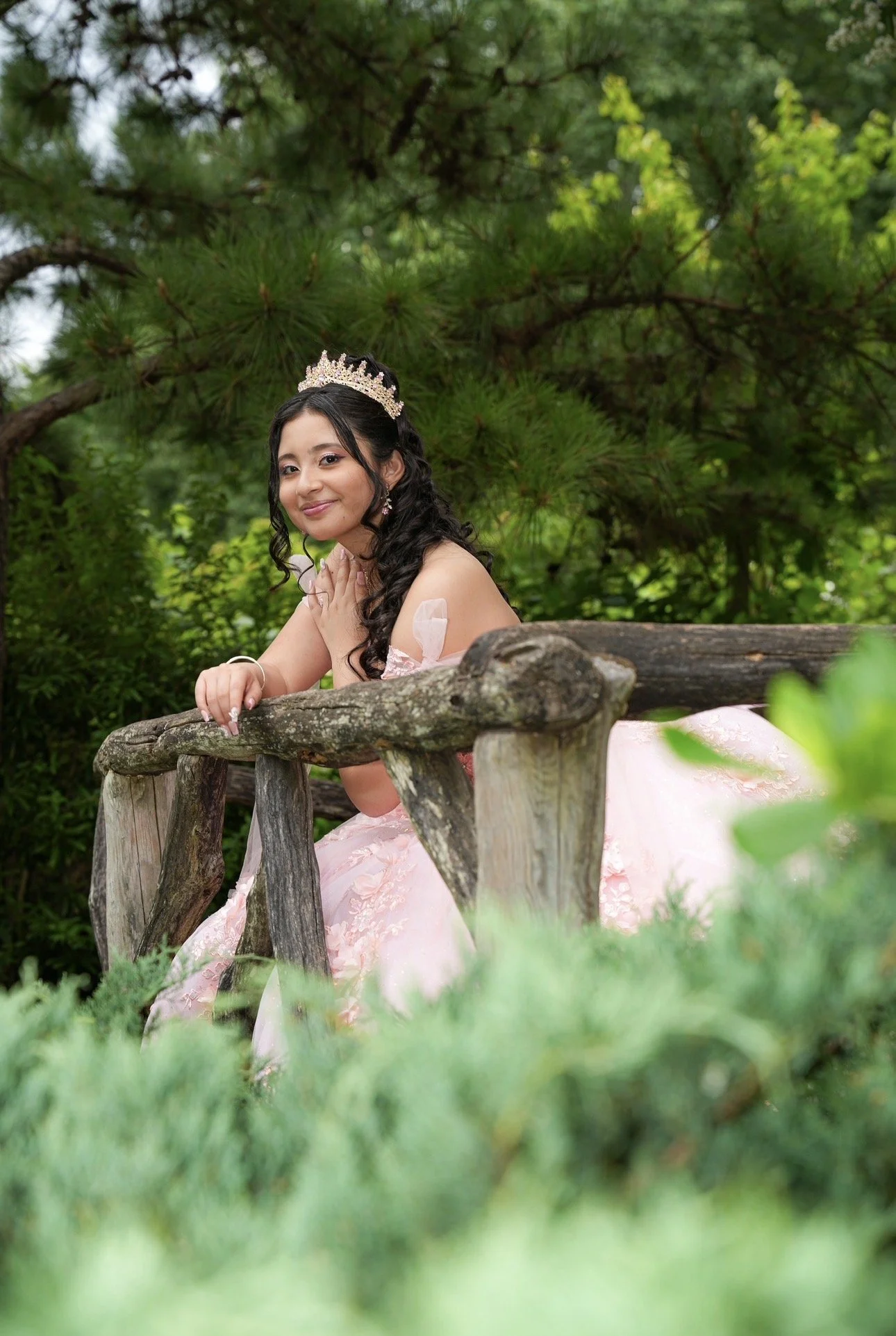Young woman in a pink dress with a tiara, sitting on a rustic wooden bridge in a lush, green outdoor setting.