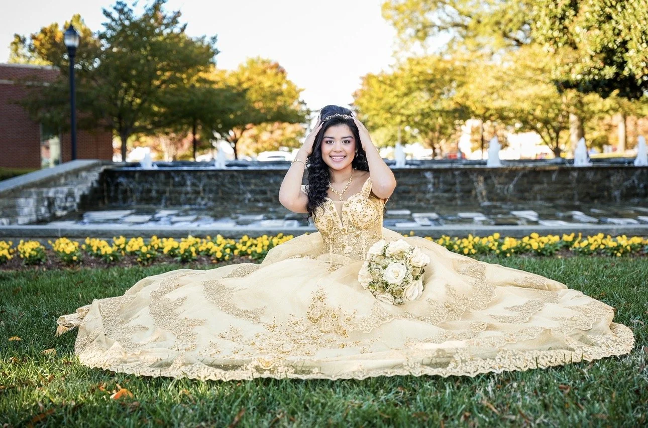 Young woman in a yellow gown sitting on grass in front of a water fountain with yellow flowers and trees in the background, smiling and holding her hair.