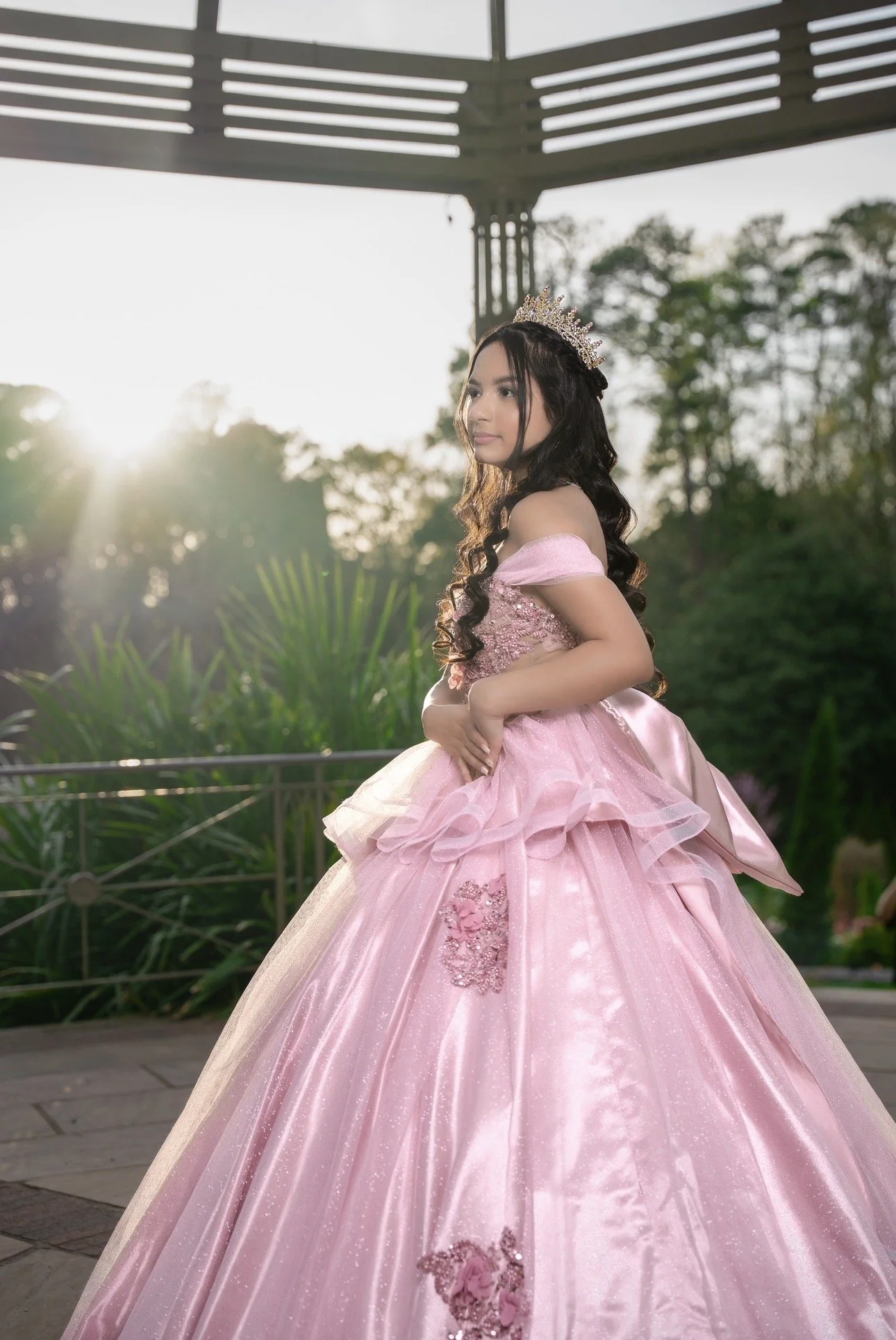 A young girl dressed in a pink ball gown with floral embroidery and a tiara, standing outdoors at sunset with trees and greenery in the background.