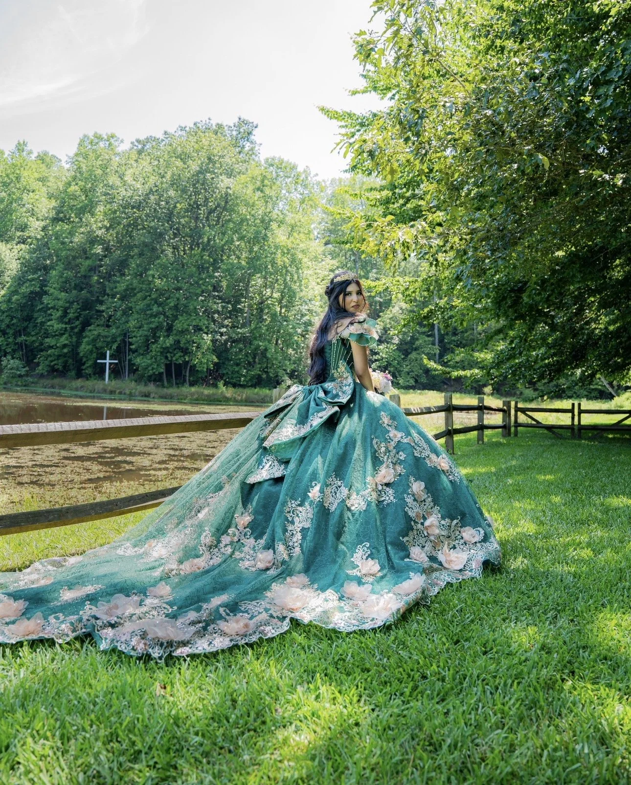 A woman wearing an elaborate teal gown with floral details standing on a grassy area near a wooden fence in a park, with trees and a pond in the background.