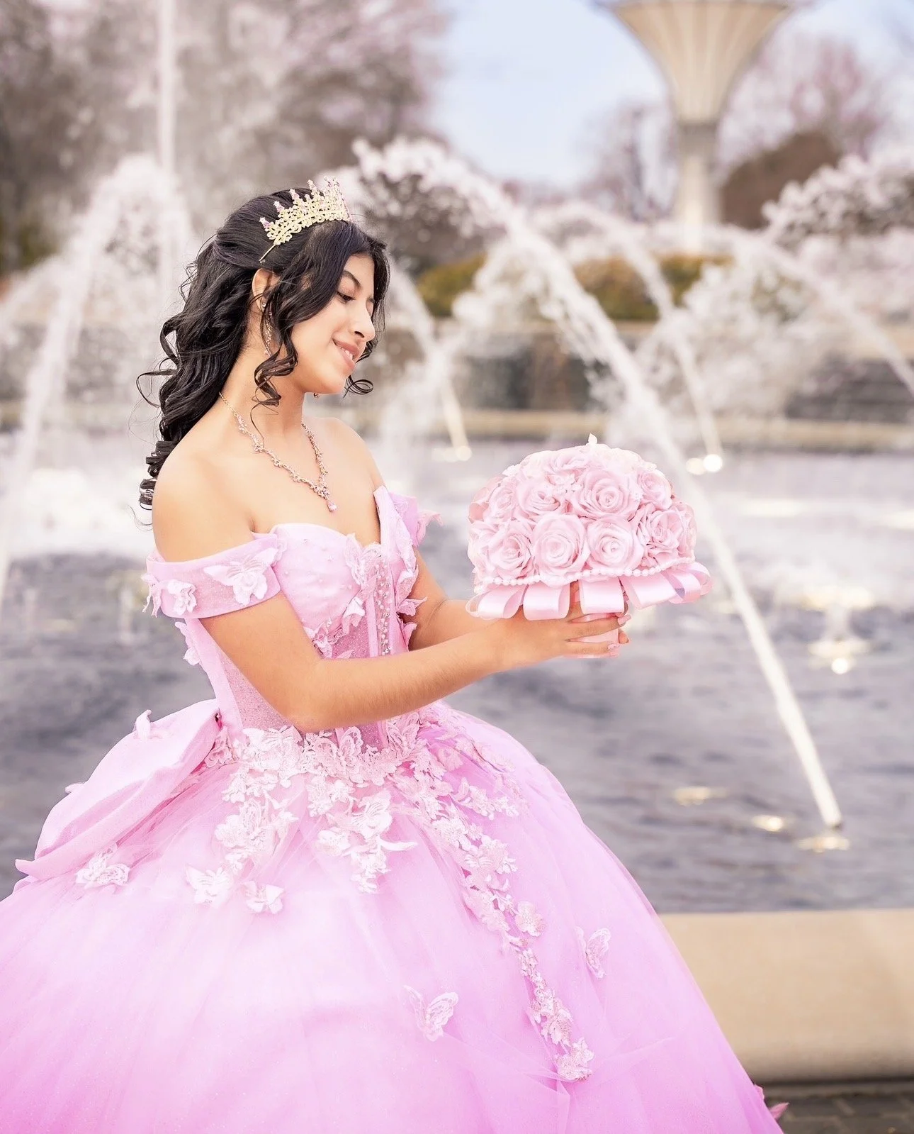 A woman in a pink, off-the-shoulder gown with lace and floral embroidery, wearing a tiara, holding a bouquet of pink roses, standing in front of a fountain with water jets.