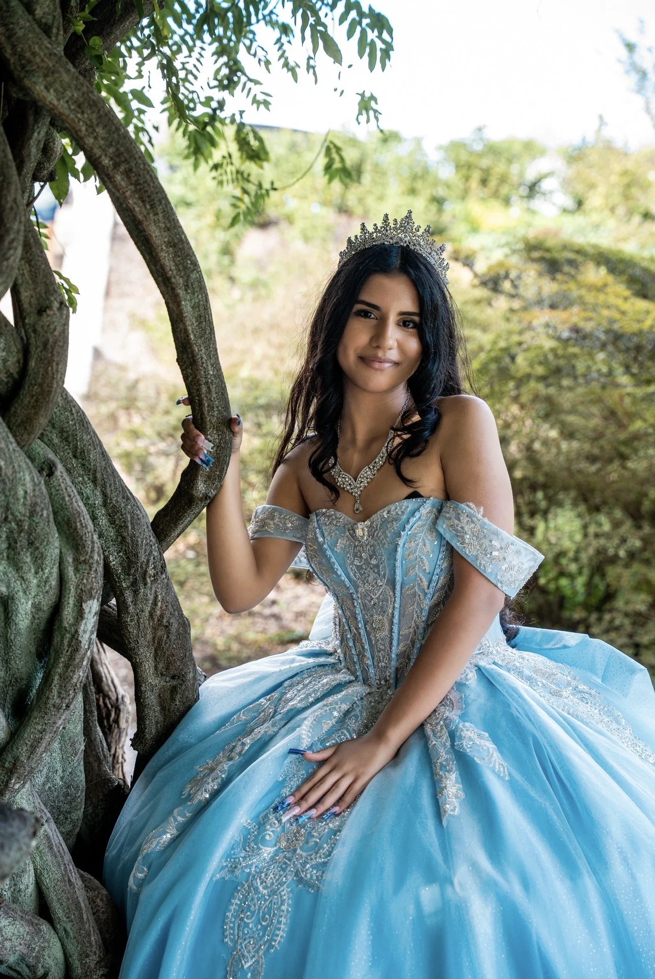 Young woman in a blue ball gown with silver embroidery and a tiara, sitting outdoors near a tree with green foliage in the background.