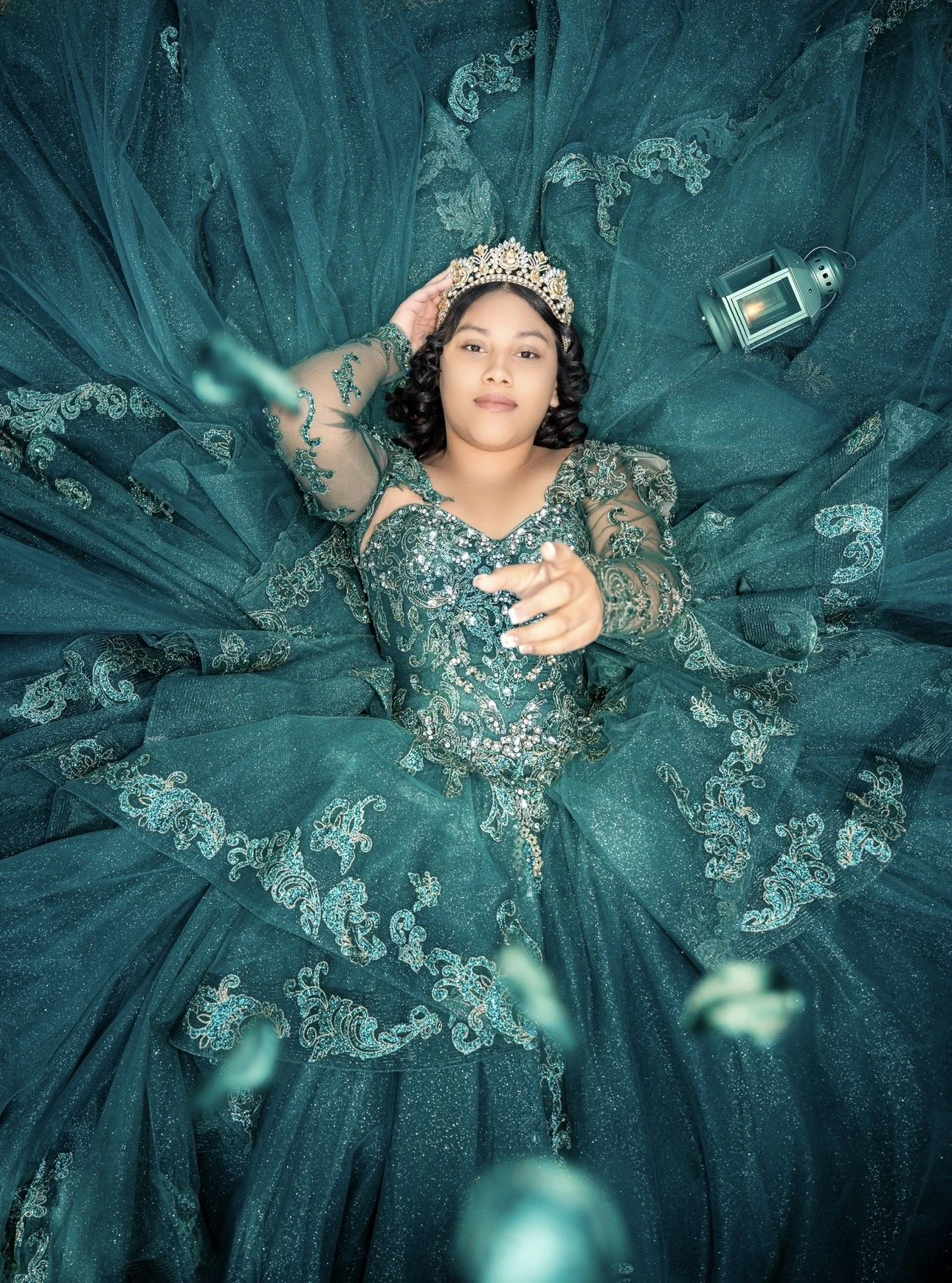 A young woman with curly hair lying on a teal dress, wearing a tiara, surrounded by teal fabric and a lantern.