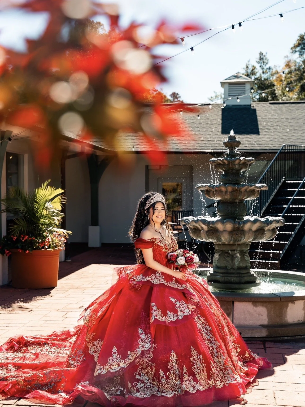 A young woman in a red quinceañera dress with white embroidery sitting near a fountain in a courtyard, holding a bouquet of flowers, with trees and string lights in the background.