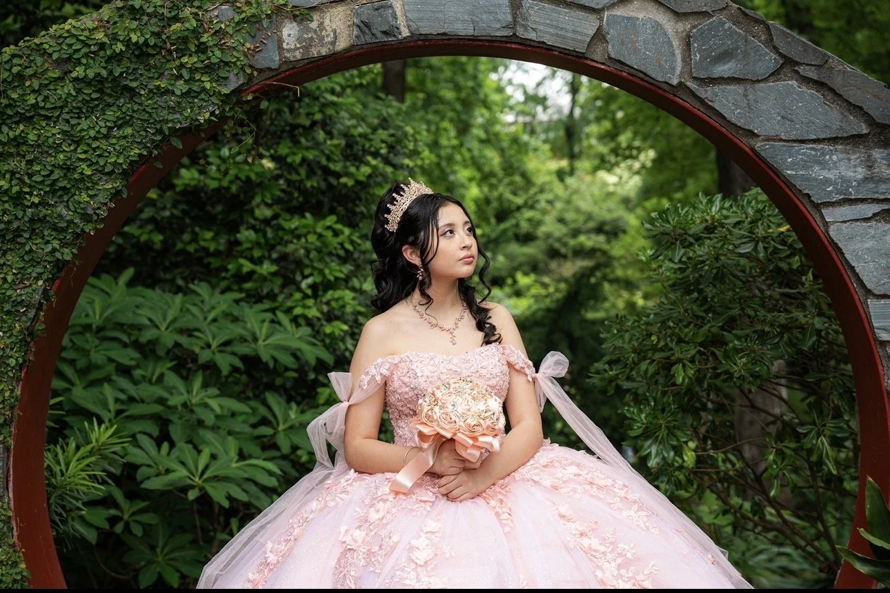 A young woman in a pink ball gown with floral details sitting outdoors inside a stone and metal circular arch, holding a bouquet of roses, wearing a tiara, with green foliage and trees in the background.