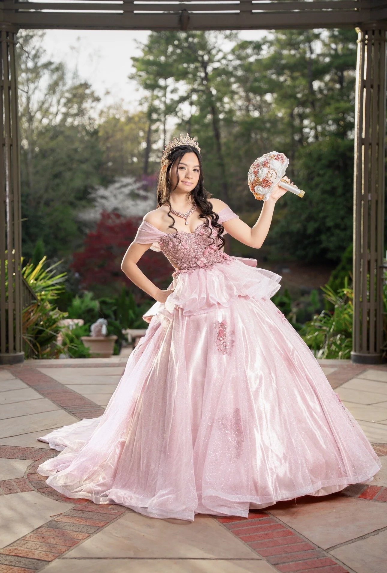 A young woman in a pink ball gown with lace and floral embellishments, wearing a tiara and necklace, holding a bouquet of roses, standing outdoors in a garden setting.