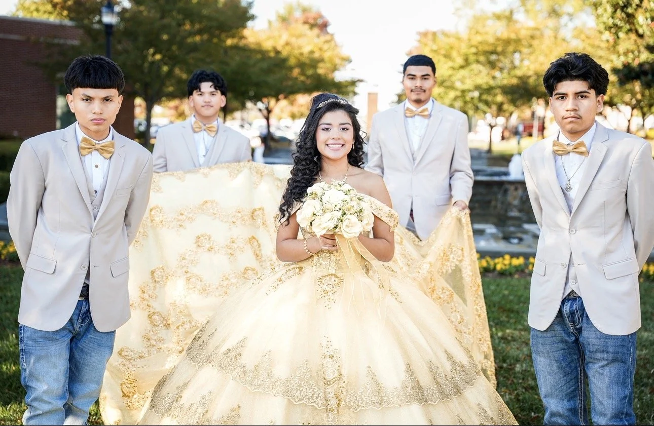 Six young people dressed in formal attire outdoors during daytime. The girl in the center wears a gold gown and holds a bouquet of white flowers. Four boys in matching light-colored suits with gold bow ties stand around her, with two boys on each sid