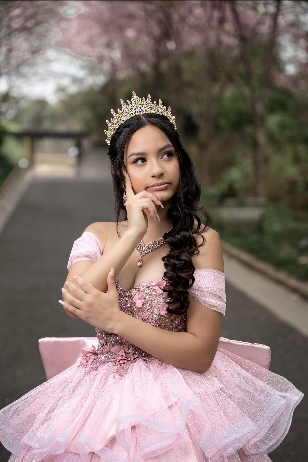 Young woman wearing a pink gown and a tiara, standing outdoors with pink blossoming trees in the background.
