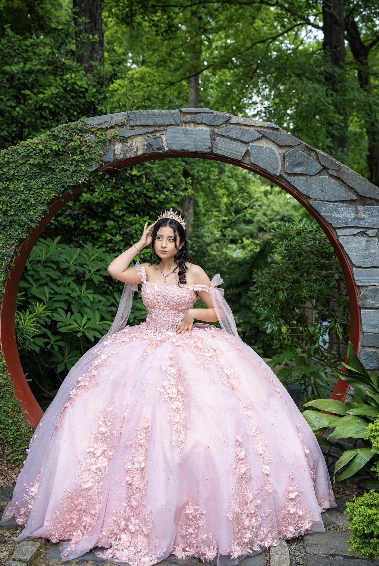 A young woman in a pink ball gown with intricate floral embroidery, sitting in a lush garden surrounded by greenery, while wearing a tiara. She is adjusting her tiara with one hand and resting the other hand on her waist.