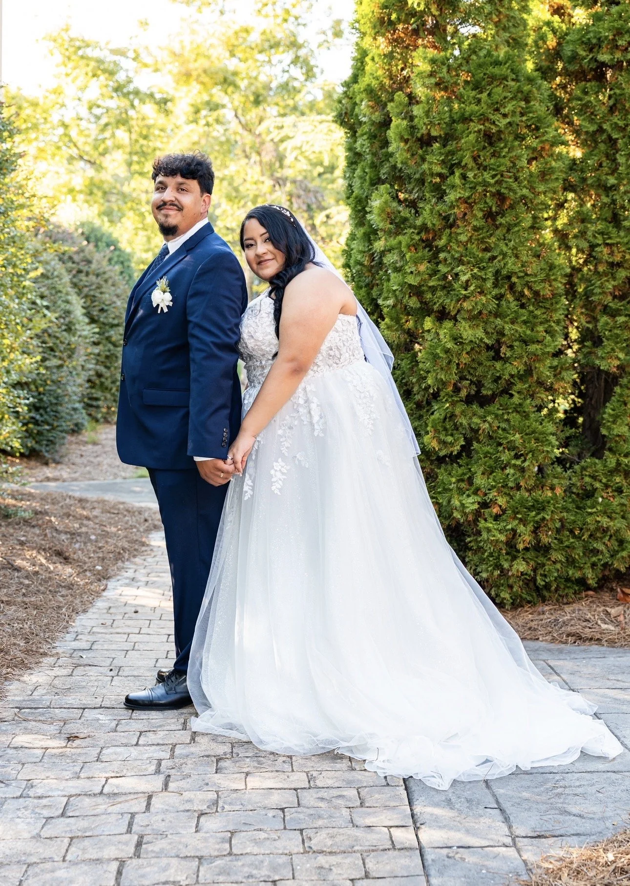 A bride and groom standing hand in hand outdoors on a paved pathway surrounded by greenery, with the bride in a white lace wedding gown and the groom in a blue suit, both smiling.