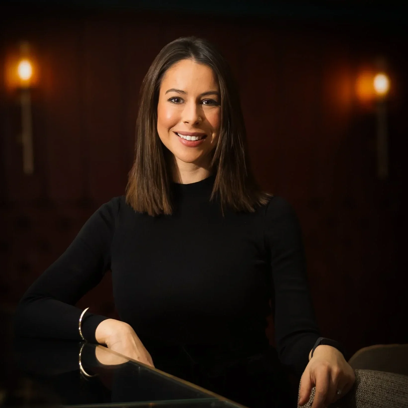 A woman with shoulder-length brown hair and a ring on her left wrist, smiling while playing a grand piano in a dimly lit setting with warm light fixtures in the background.
