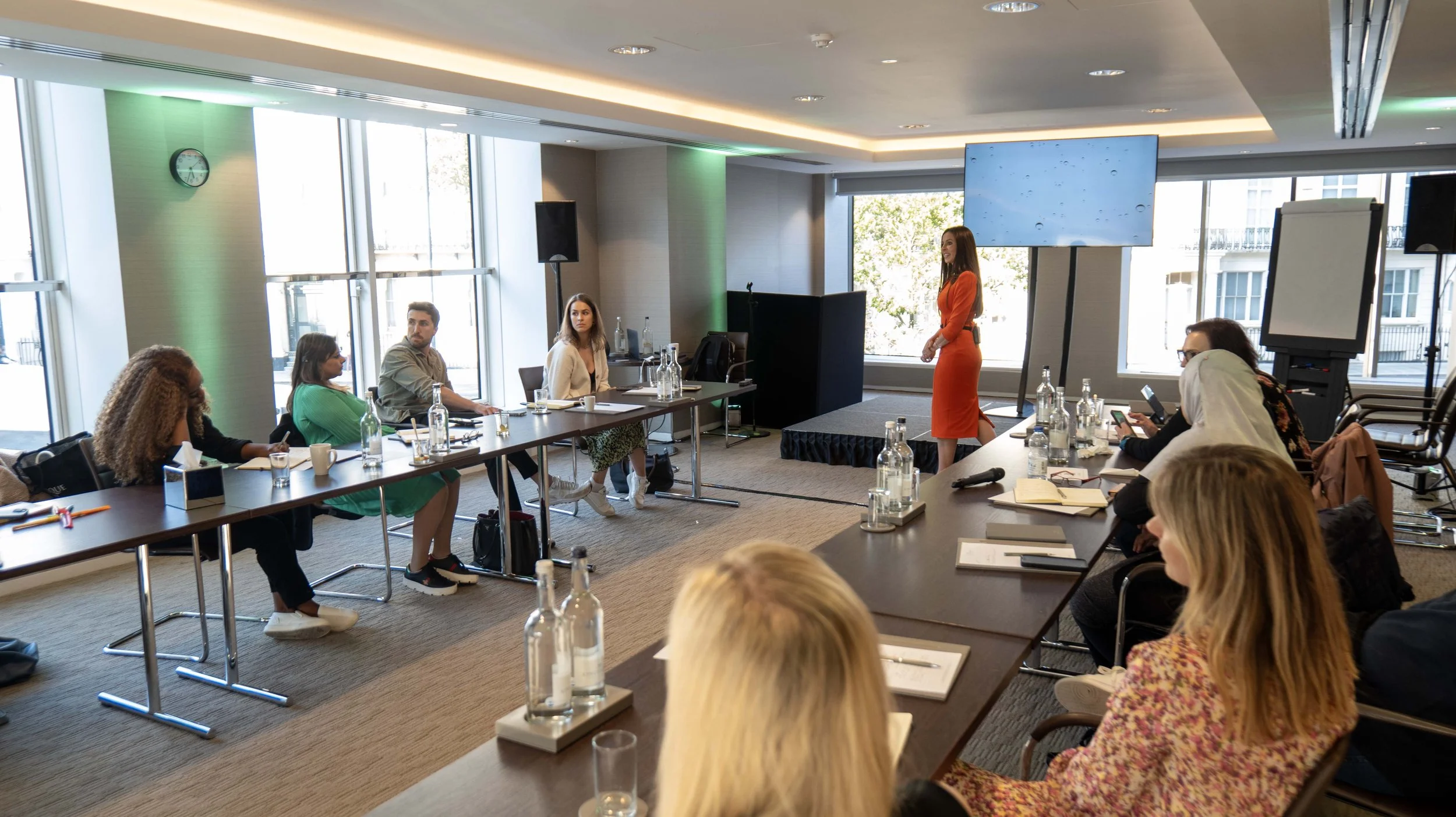 A woman in an orange dress is giving a presentation in a conference room with attendees seated at tables.