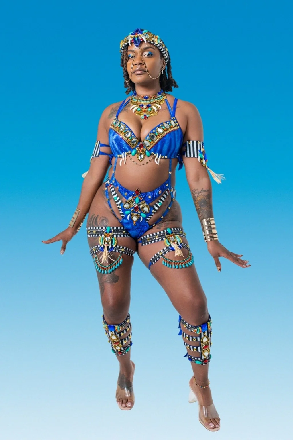 Woman wearing a vibrant blue costume decorated with beads, sequins, and jewelry, standing against a plain blue background.