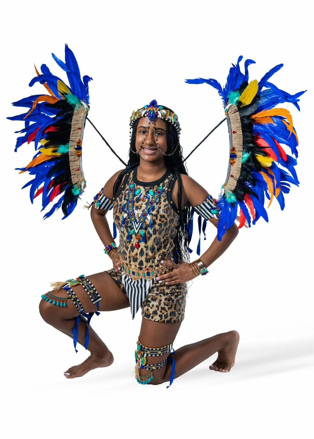 A woman in elaborate, colorful tribal costume with large feathered wings, jewelry, and animal print clothing, kneeling on one knee with a serious expression.